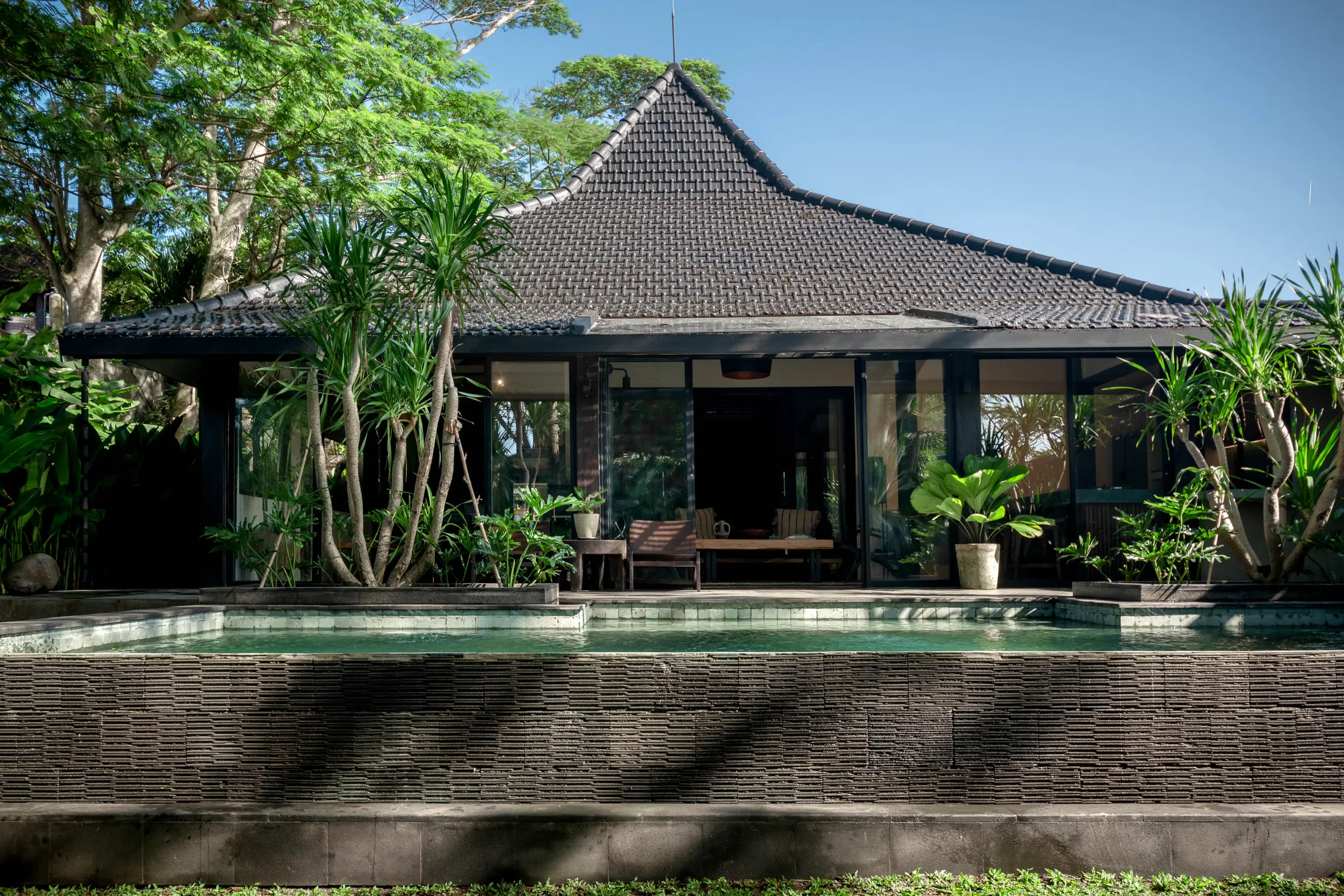 Single-story villa with a tall dark-tiled pyramid roof behind a raised pool and dark woven-tile retaining wall, framed by tropical palms – The Lo-Fi Villa, a 3-bedroom villa in Cemagi, Bali