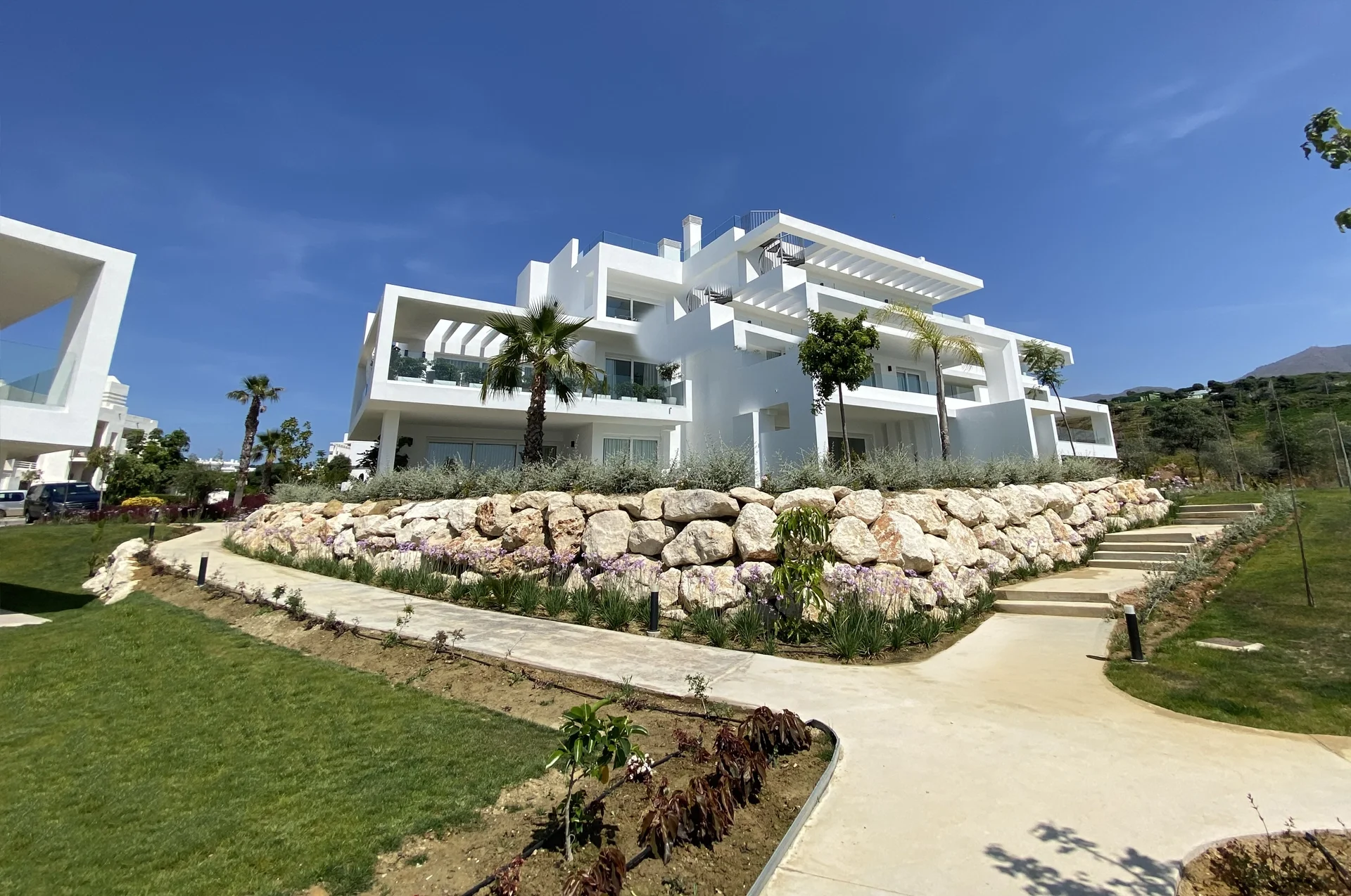 Photo of a white cubist Mediterranean-style villa building with palm trees behind a boulder retaining wall with purple flowering plants and a curved footpath – Alcazaba Lagoon IV, a 3-bedroom villa in La Alcazaba Nº312, Cortijo El Beneficiado, Casares, Málaga