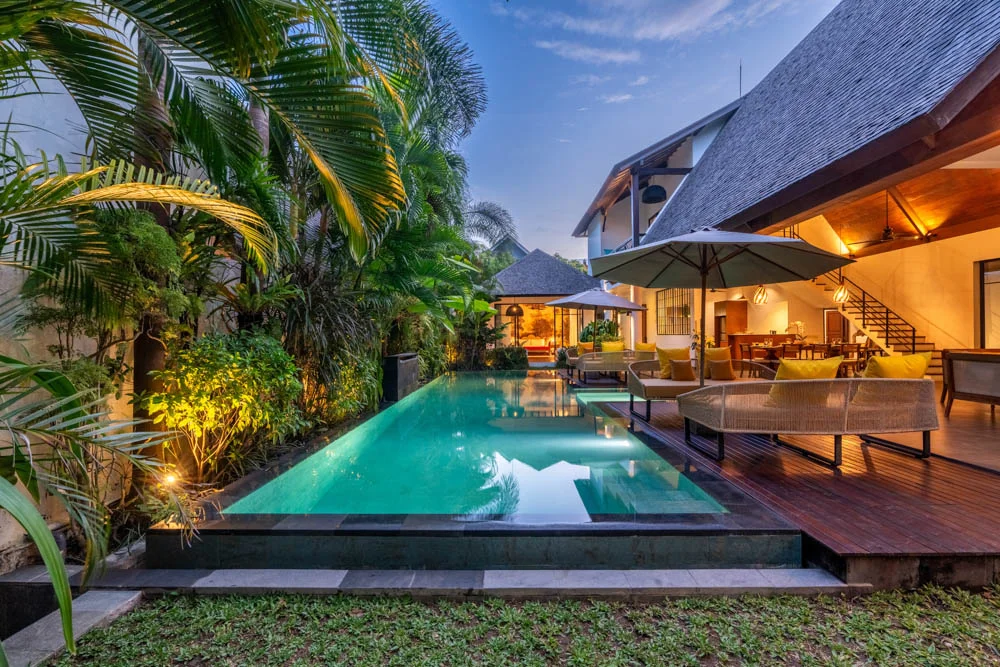 Twilight view of a tiled-roof pavilion with a sunken lap pool, rattan lounge seating with yellow cushions and a white umbrella on a wood deck – Villa Soka, a 4-bedroom villa in Pererenan, Bali