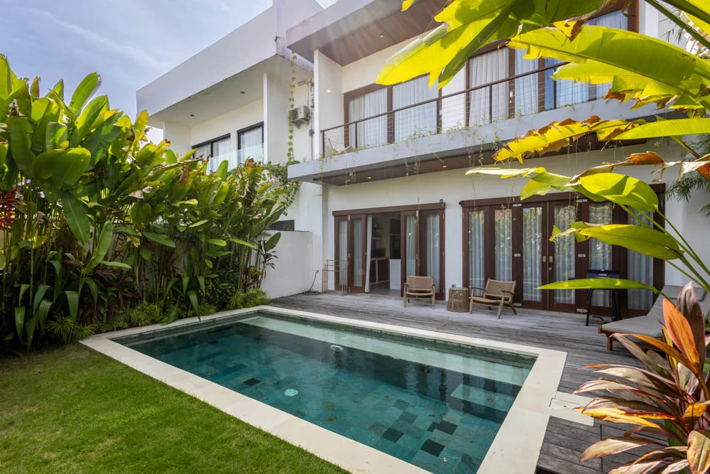 Two-story white villa with dark wood-framed doors and a cable-railed balcony, beside a rectangular pool set into a wood deck – Villa Lula, a 3-bedroom villa in Pererenan, Bali