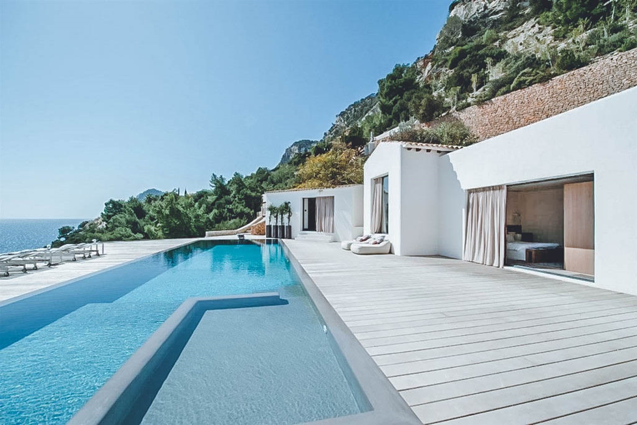 Photo of a white villa built into a rocky cliffside, with an L-shaped infinity pool along a wooden deck overlooking the sea – Villa Bailey, a 6-bedroom villa in Es Cubells