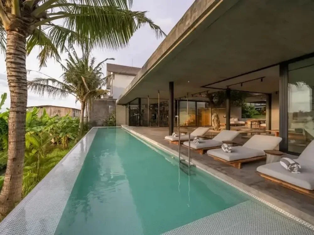 Concrete-roofed villa with a long narrow lap pool running past a glass facade, lined with cushioned wooden sun loungers and shaded by tall palm trees – Villa Mozart, a 5-bedroom villa in Kayu Tulang, Canggu, Bali