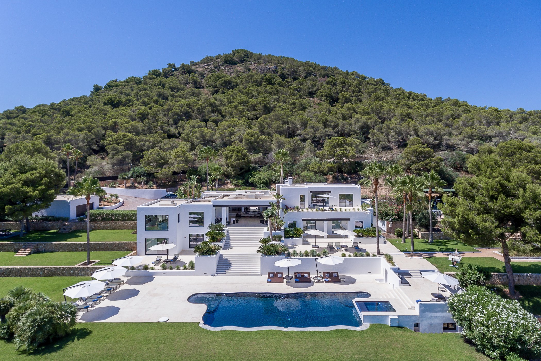 Aerial daytime view of a large white modern villa below a pine-covered hillside, with a free-form pool, sunken spa corner and terraced lawns – Can Nemo, a 6-bedroom villa