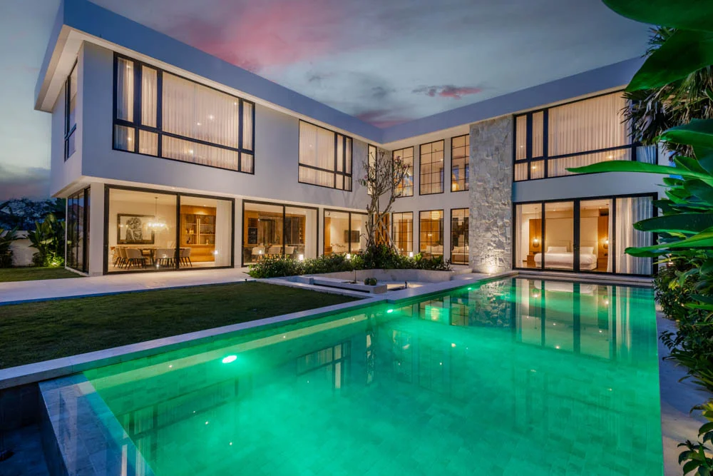 Two-story white L-shaped villa with black-framed windows lit from inside at dusk, wrapping around a large green-lit pool with a stone accent column – Villa Athena, a 5-bedroom villa in Bumbak, Umalas, Bali
