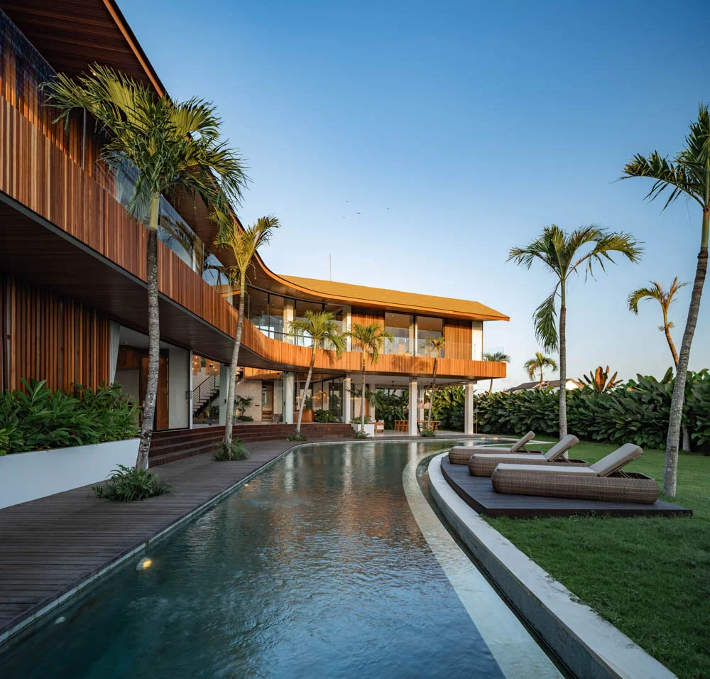 Two-story timber-clad villa with a sweeping curved balcony following a curving pool, lined with palm trees and sun loungers on the lawn – Villa Efiren M2-M3, a 5-bedroom villa in Tumbak Bayuh, Pererenan, Bali