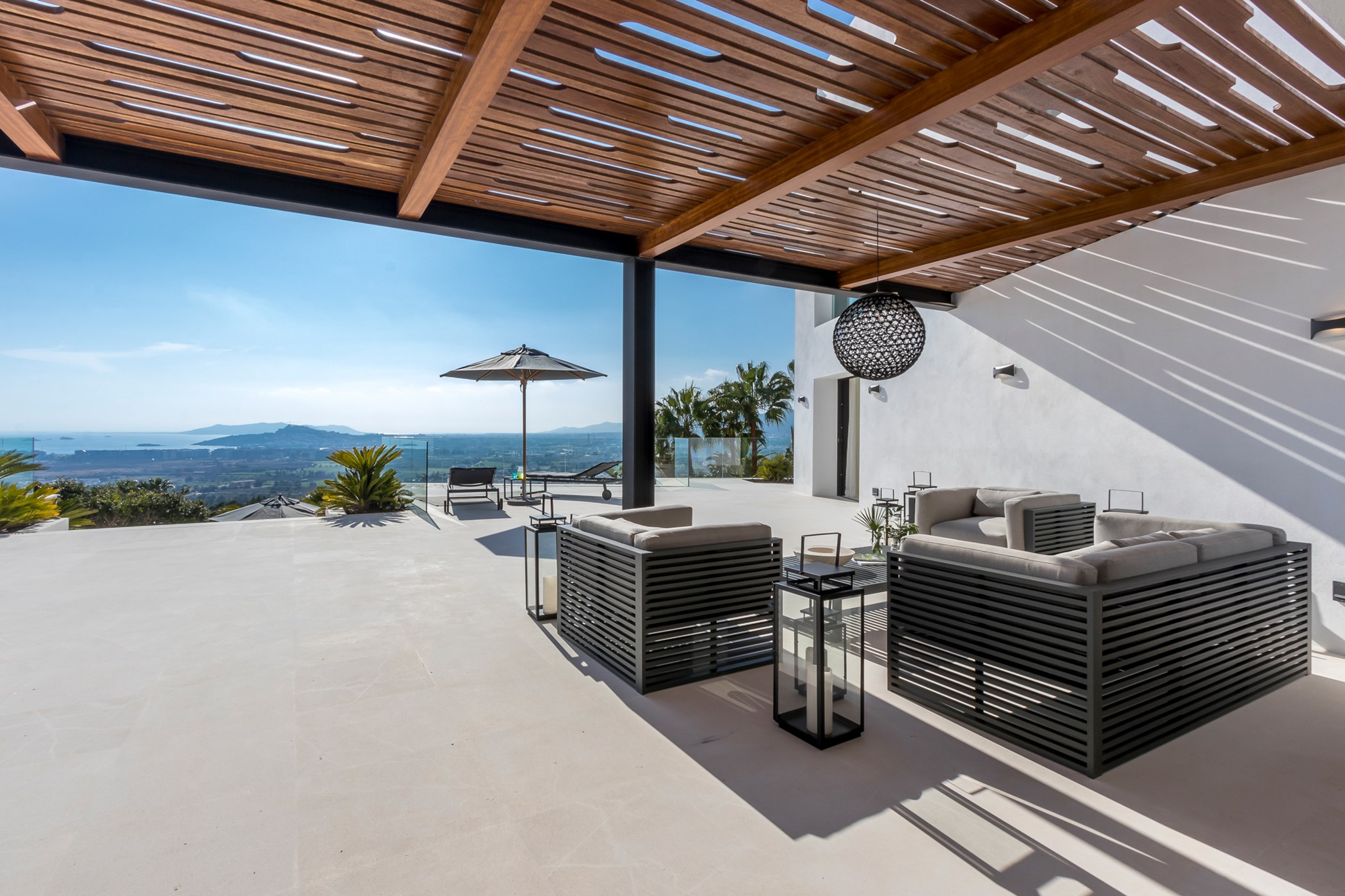 Photo of a covered terrace with a wood-slat pergola, dark slatted outdoor sofas and lanterns overlooking a panoramic coastal view – Villa Shiva, a 12-bedroom villa in Can Rimbau