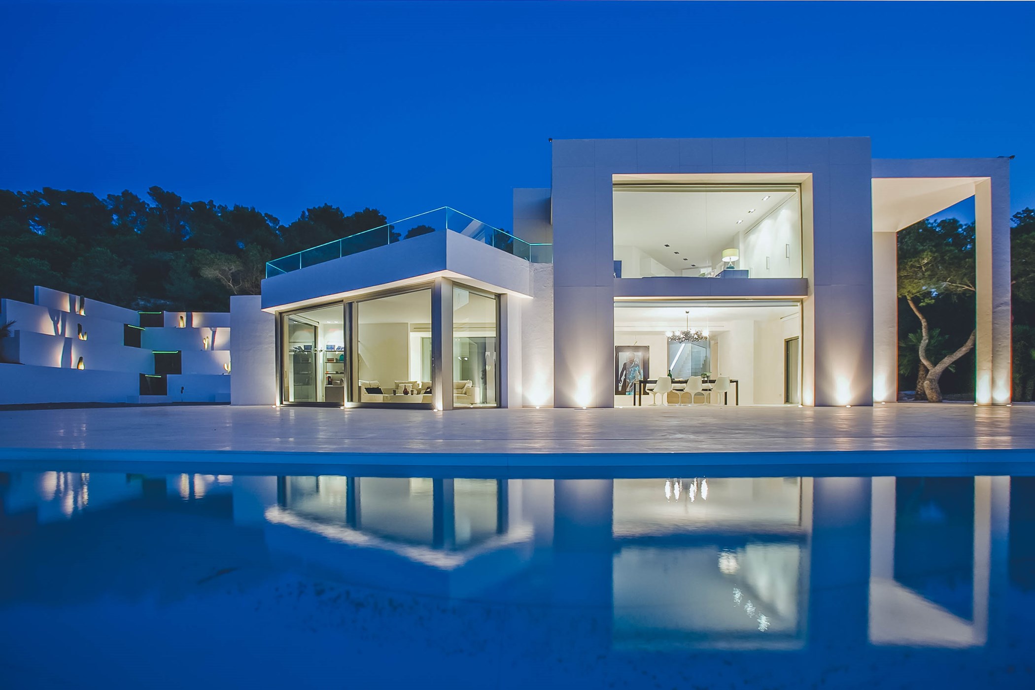 Twilight photo of an angular white cubist villa with illuminated glass-walled interiors and a sculptural relief wall, reflected in a still pool – Villa Adalyn, a 6-bedroom villa in Sant Josep