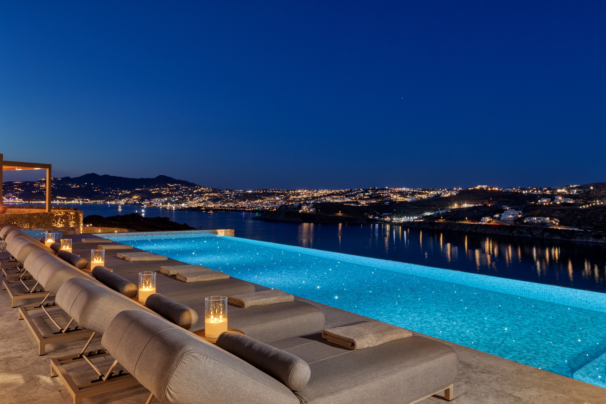 Infinity pool terrace with loungers and candle lanterns at night, overlooking a lit coastal bay and hillside town in the distance – Clio Estate, a 7-bedroom villa