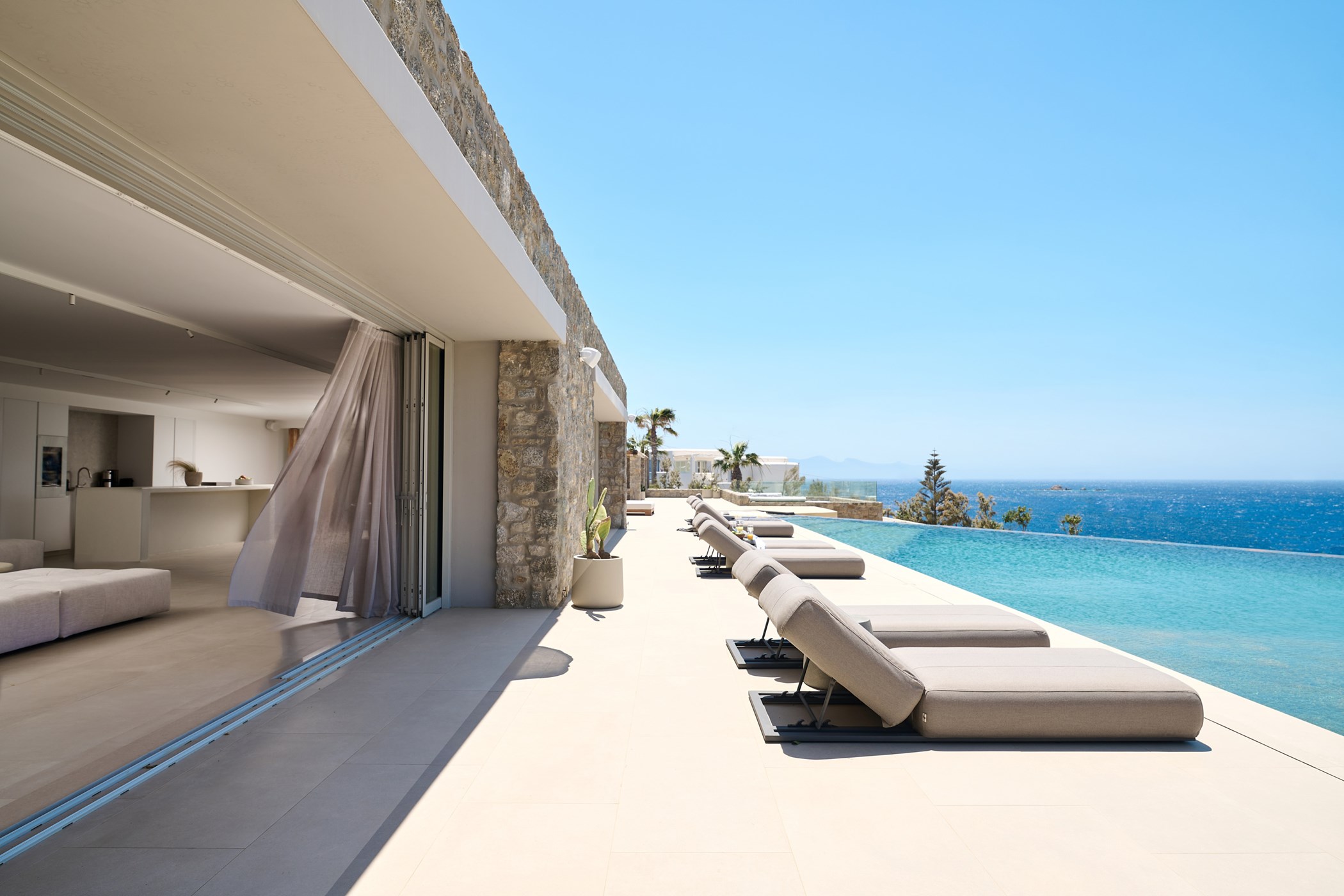 Stone-clad villa terrace with a row of sunloungers along an infinity pool overlooking the sea, open living space visible through sliding doors – Blue Sky, a 9-bedroom villa