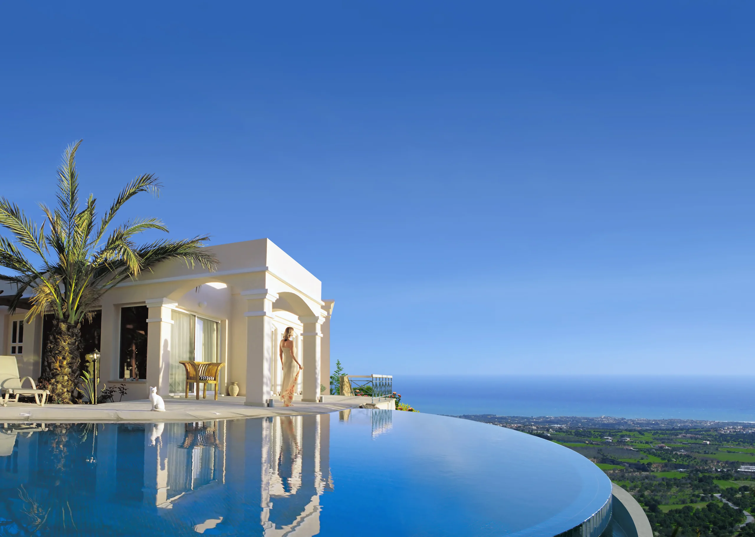 Cream-colored villa with a columned porch and palm tree beside a curved infinity pool overlooking a green countryside panorama toward the sea – Kamares Village, a 3-bedroom villa in Kamares, Paphos, Cyprus