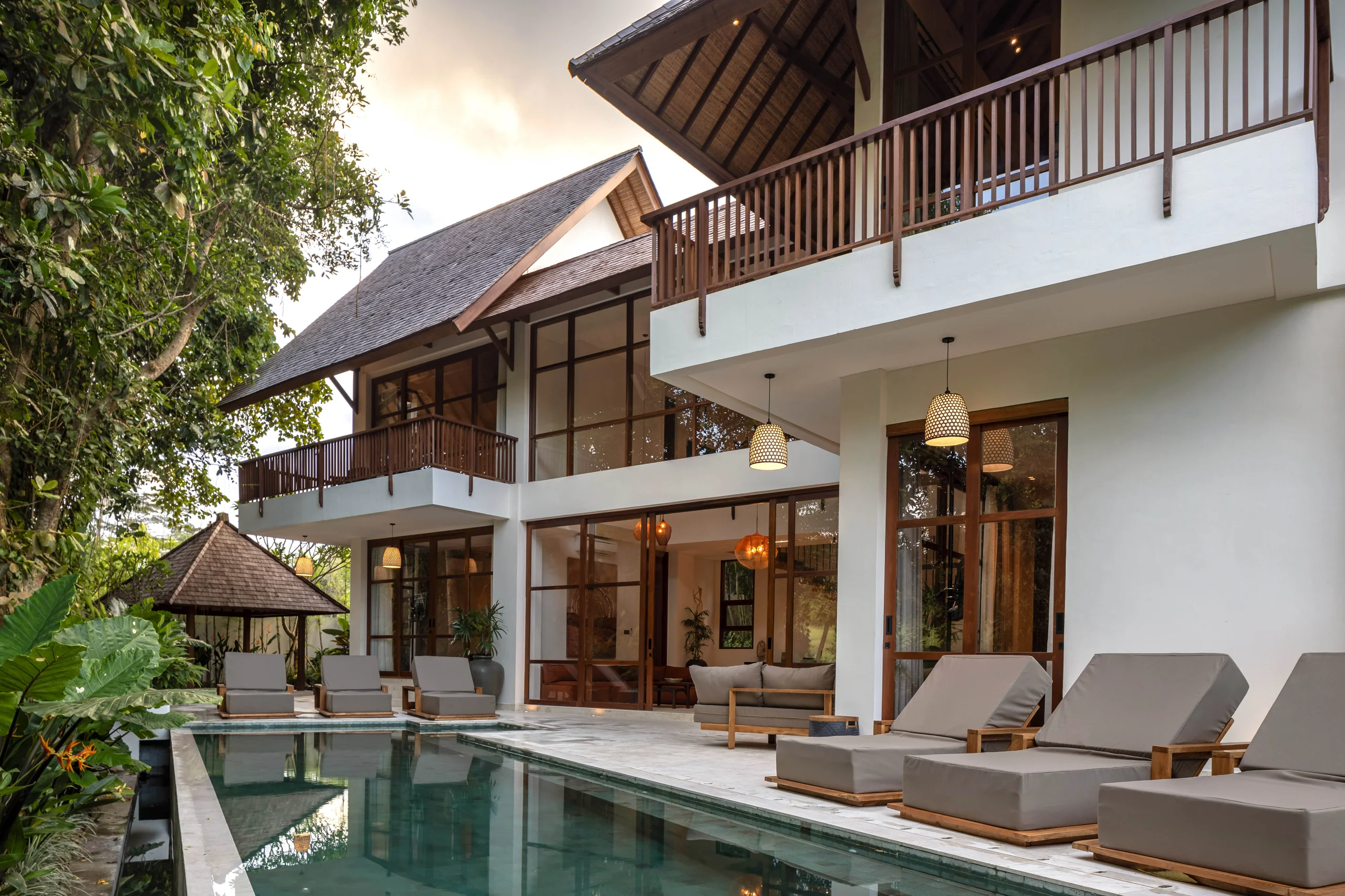 Two-story villa with a steep shingle roof and wood-railed balconies, a separate round-roofed gazebo, and a long pool with wood lounge chairs at dusk – Villa Norton, a 5-bedroom villa in Nyanyi, Tabanan, Bali