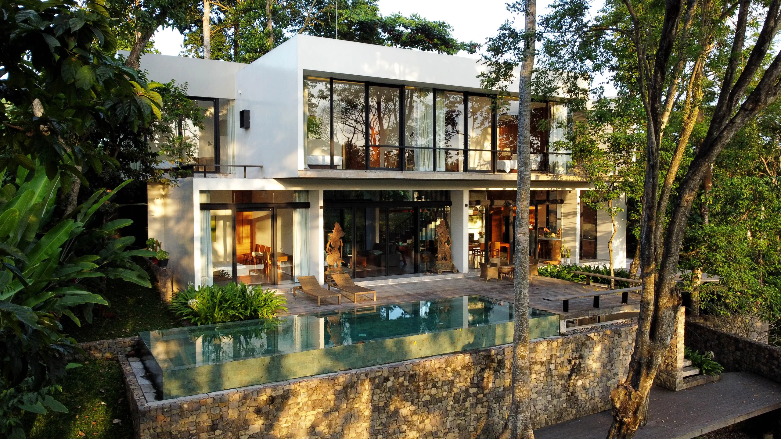 White cubic modern villa set among dense trees, with floor-to-ceiling glass walls and an elevated infinity pool edged by a stone retaining wall – Villa Maxey, a 4-bedroom villa in Buwit, Bali