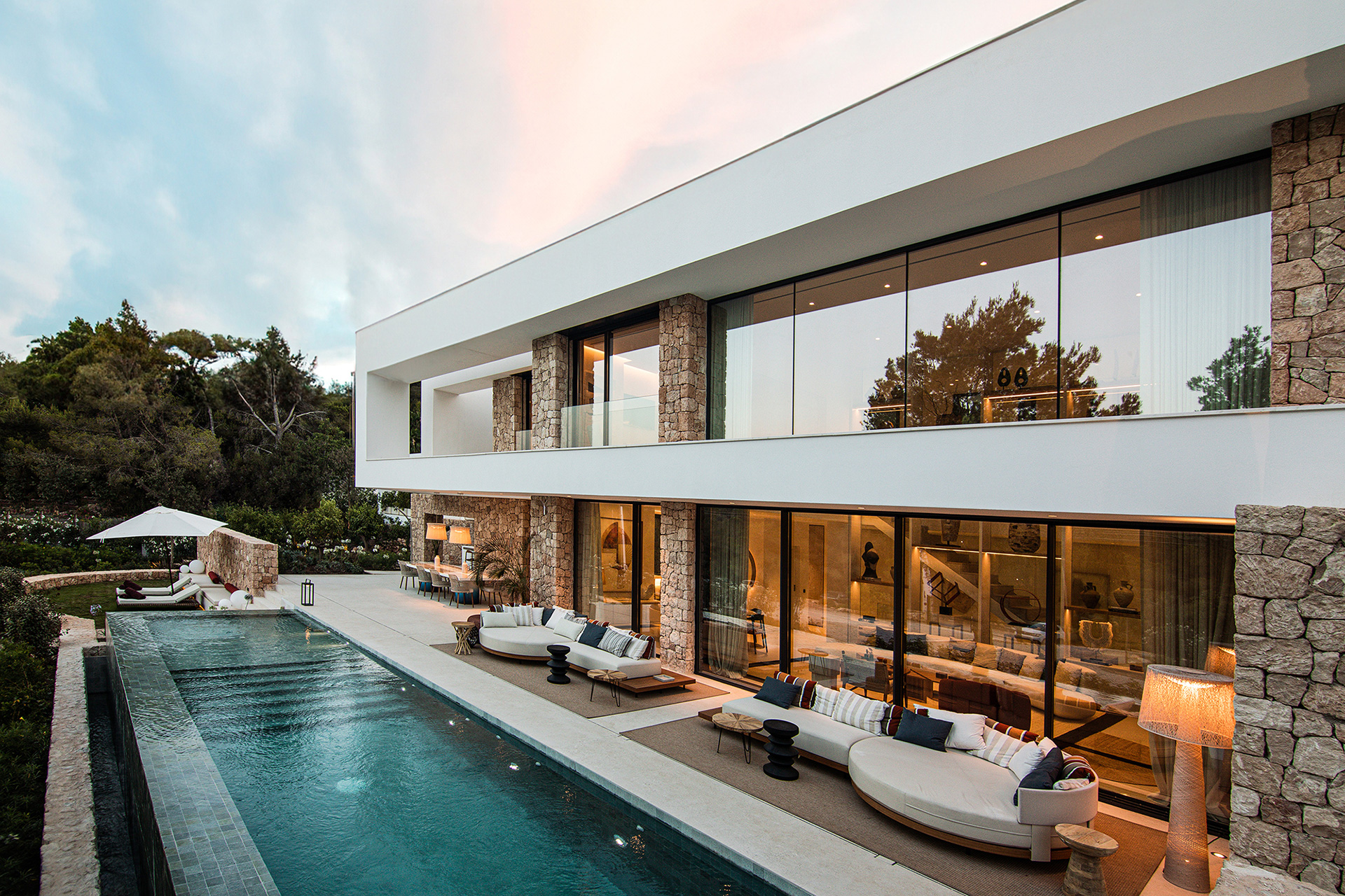 Two-story cantilevered villa with stone-clad columns and glass walls behind curved outdoor sofas on a terrace beside a long infinity pool – Corallisa, a 5-bedroom villa in Roca Llisa, Eivissa, Illes Balears