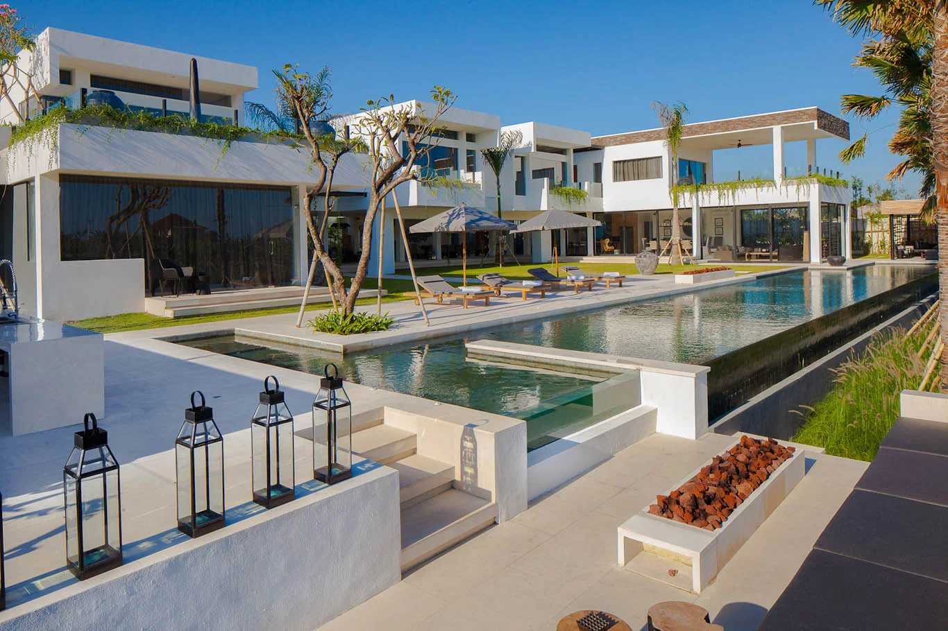 Sprawling white modern villa complex around a long dark-tiled infinity pool bordered by lawn, with lounge chairs, umbrellas and lanterns on the terrace – Villa Mevvah, a 5-bedroom villa in Tumbak Bayu, Pererenan, Bali