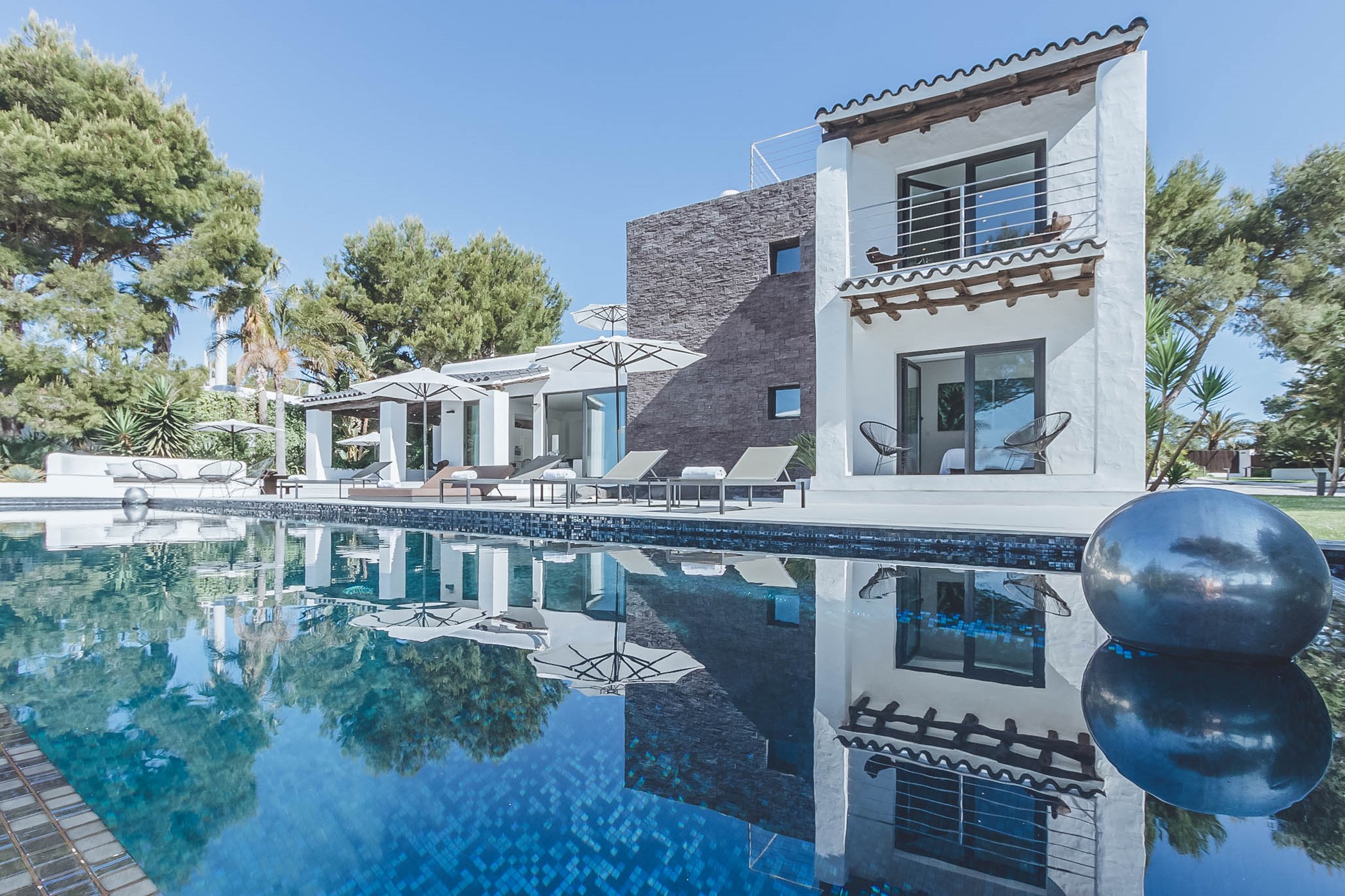 Two-story Ibiza-style white villa with a dark stone-clad tower, rustic wood-beam balcony trim and terracotta roof tiles beside a blue-tiled pool with dark spherical sculptures – Villa Vivian, a 6-bedroom villa