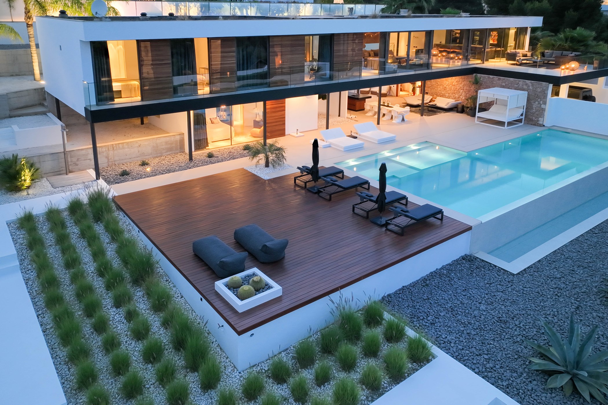 Aerial dusk view of a cantilevered white villa above a sunken wooden lounge deck with daybeds, a gravel garden and a large rectangular pool with a built-in spa – Villa Mila, a 4-bedroom villa