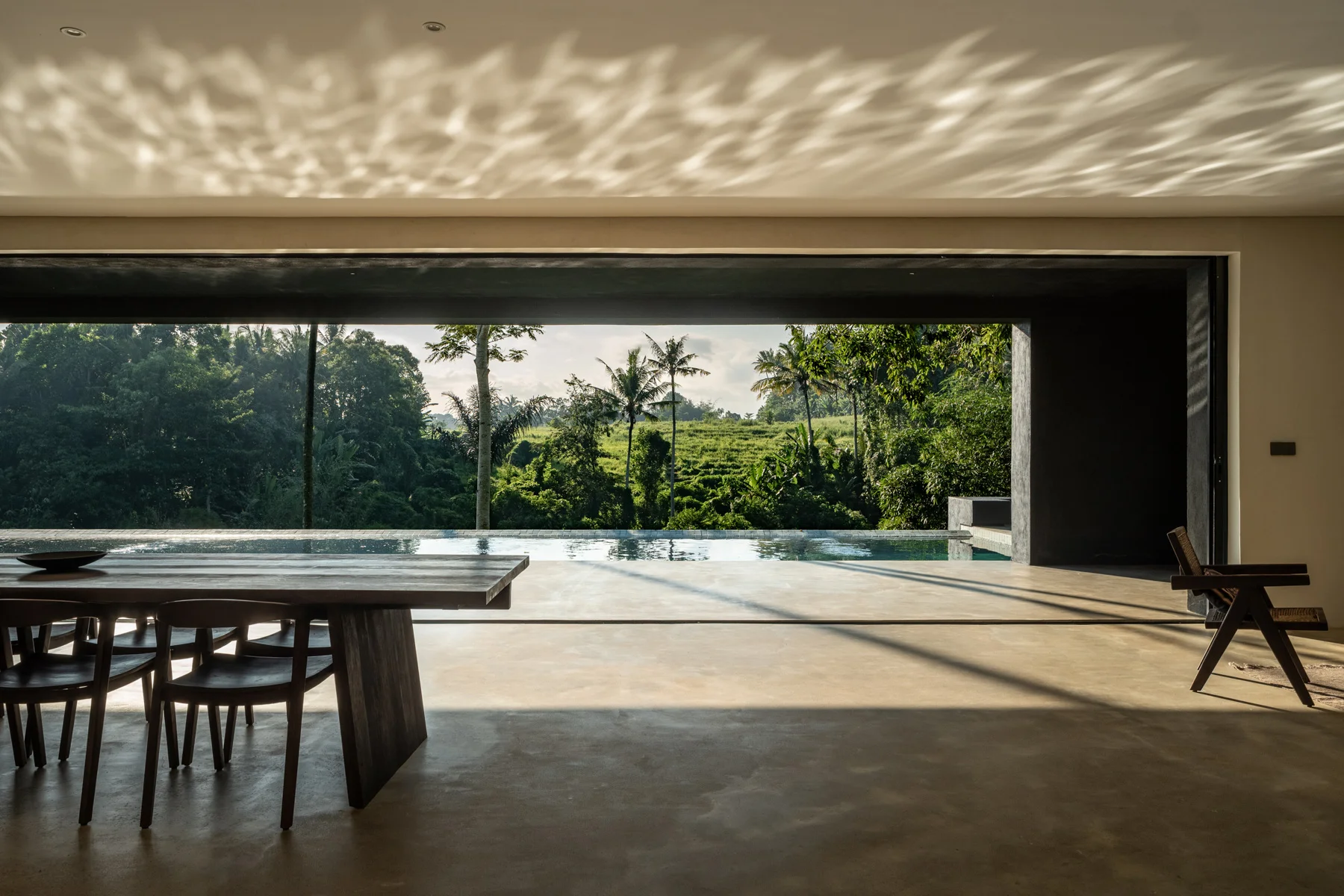 Open-air dining pavilion with a long wood table, looking out past an infinity-edge pool to dense jungle tree line at golden hour – Villa Palais, a 4-bedroom villa in Kedungu, Tabanan, Bali