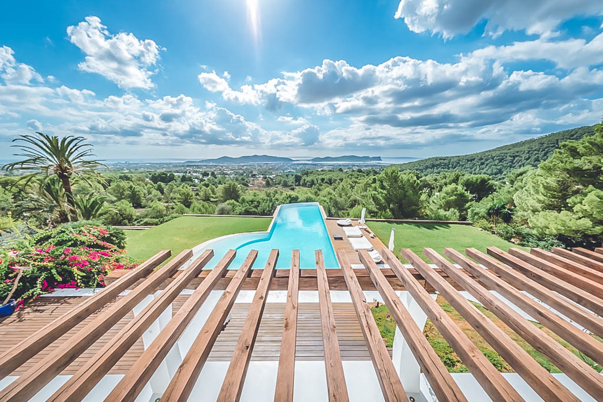View through a wooden slatted pergola over an elevated curved infinity pool and lawn toward wooded hills and the coastline in the distance – Villa Margo, a 6-bedroom villa