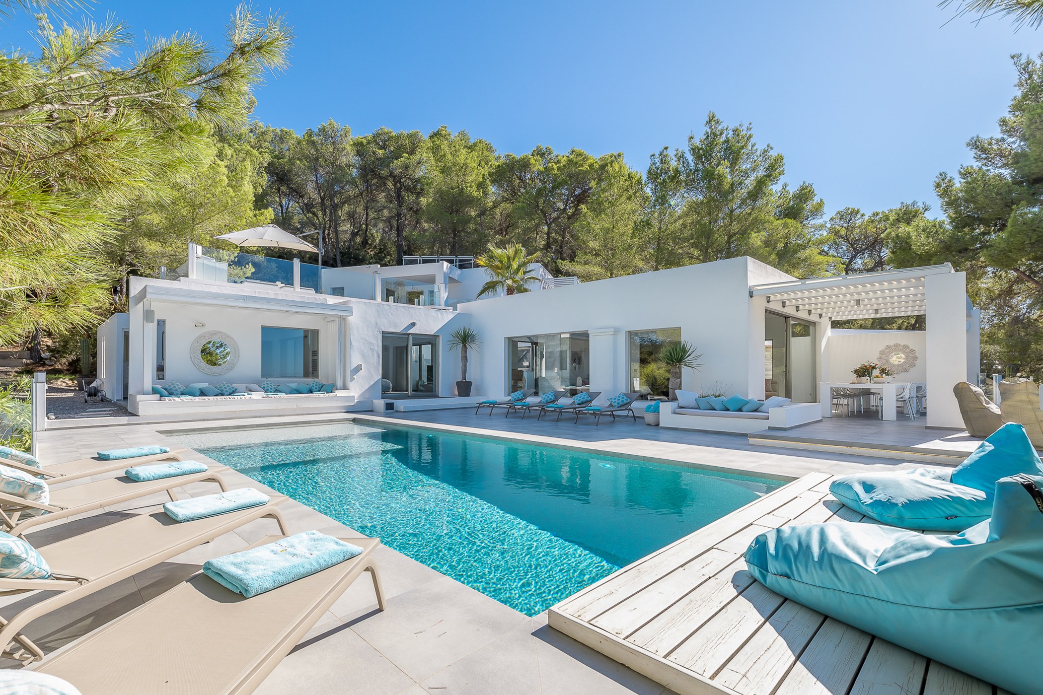 All-white Ibiza-style villa with built-in banquette seating and turquoise cushions around a rectangular pool, a pergola-shaded dining terrace and pine forest behind – Villa Ivy, a 6-bedroom villa
