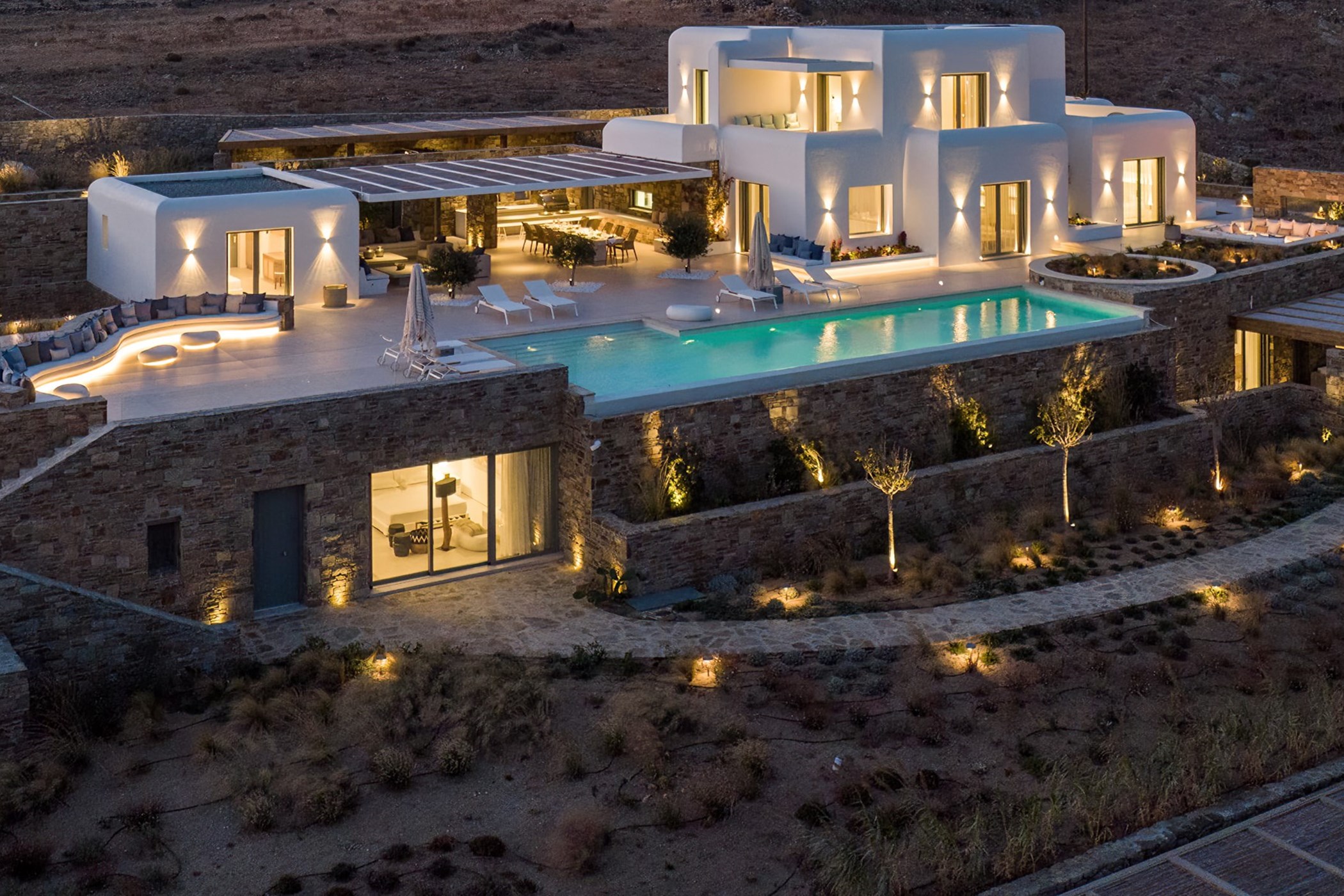 Aerial night view of a Cycladic-style white villa with cube-shaped illuminated volumes on a stone base, a lit rectangular pool and curved lounge terrace on a bare hillside – Villa Ines, an 8-bedroom villa