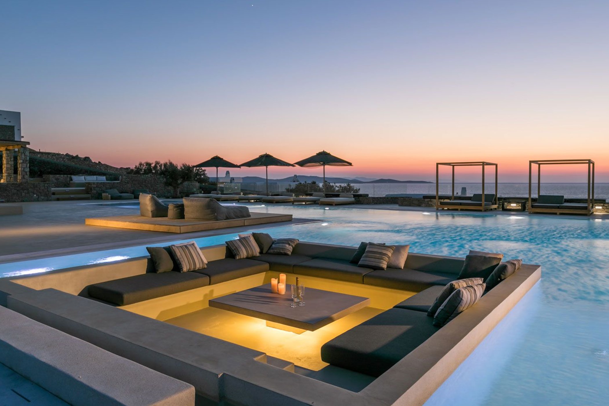 Sunken lounge island built into an infinity pool at sunset, with an illuminated corner sofa around a candlelit table and the sea and islands beyond – Villa Gaia, an 8-bedroom villa