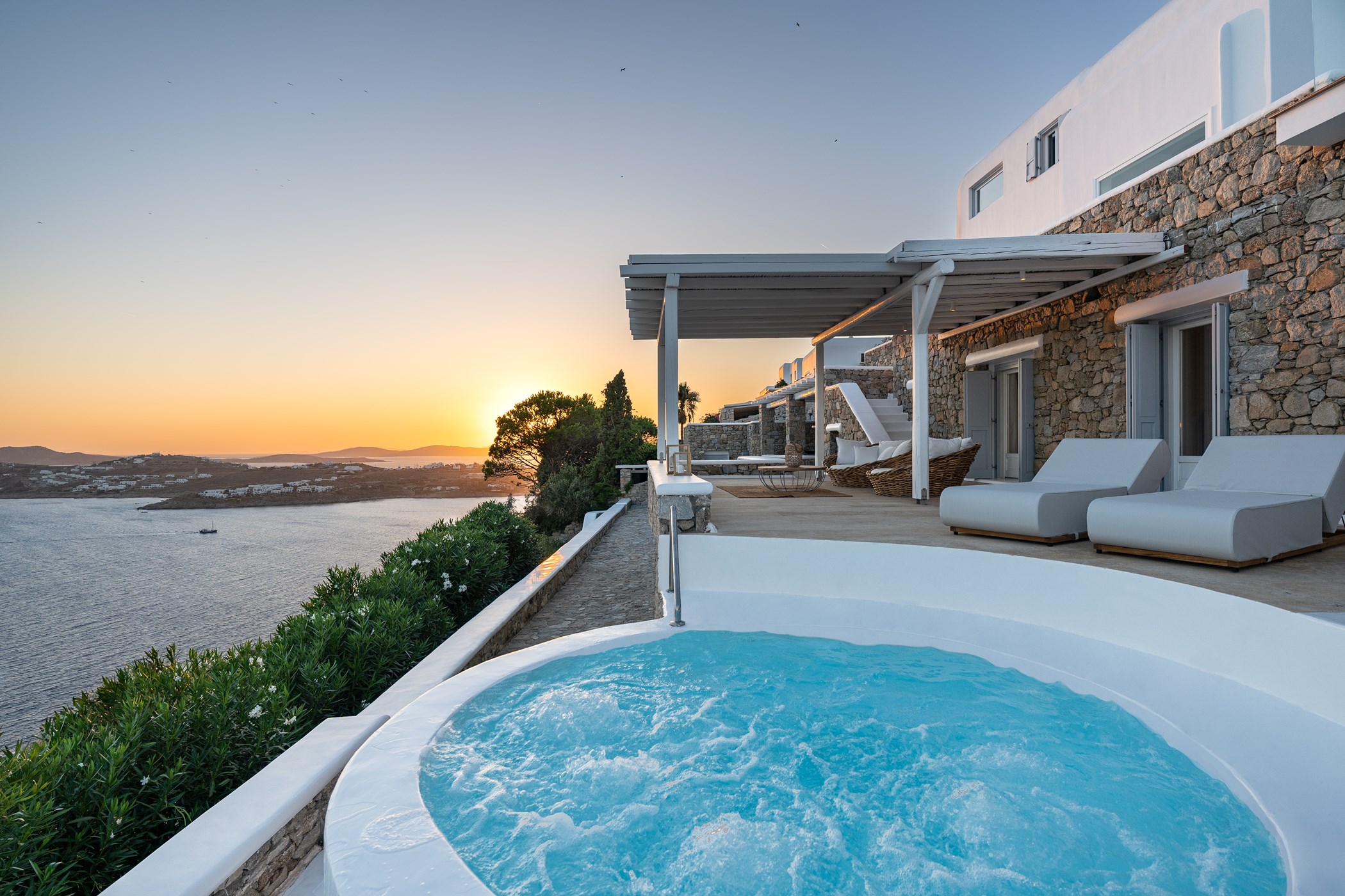Round bubbling hot tub on a clifftop terrace of a stone-clad villa at sunset, overlooking a bay, with a pergola-covered wicker lounge nearby – Villa Amy, an 8-bedroom villa