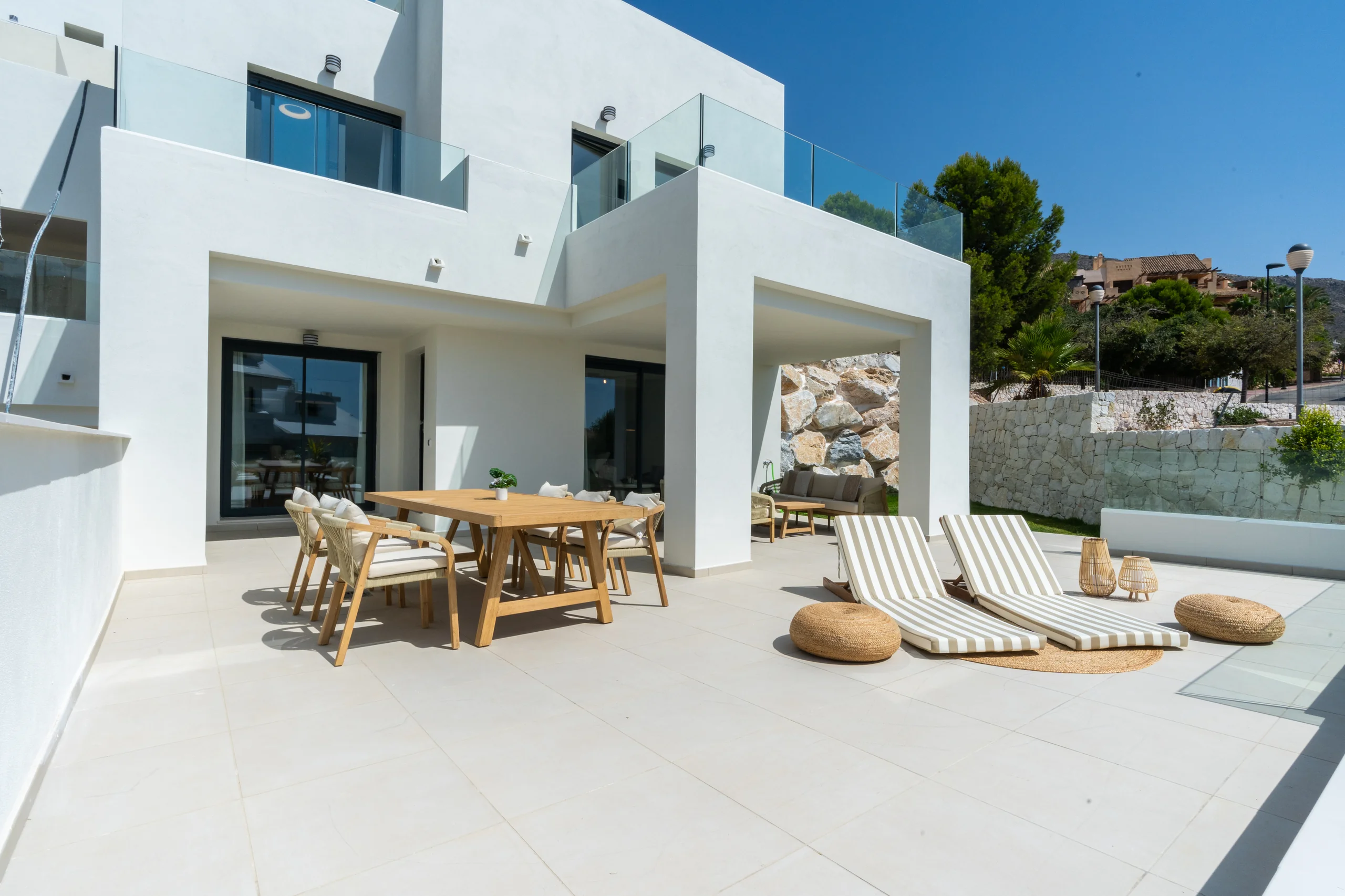 White modern villa terrace with a wooden dining table, striped sunloungers and a stone accent wall overlooking a hillside – Mane Residences, a 2-bedroom apartment in Marbella, Málaga, Costa del Sol