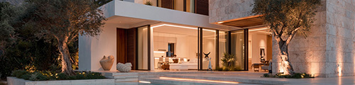Modern stone villa entrance at dusk, framed by olive trees with warm interior lighting through glass doors – Cyprus