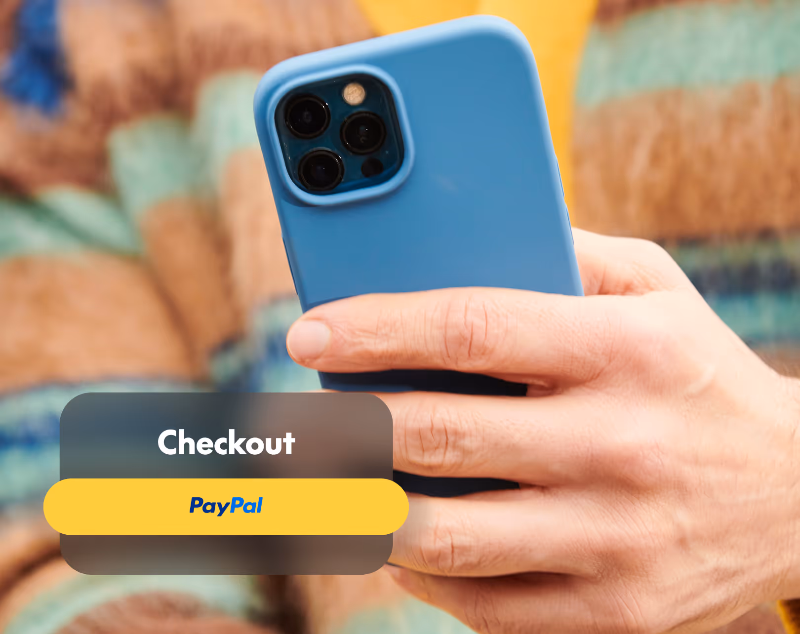 Hand holding a blue smartphone with a yellow PayPal checkout button overlay.