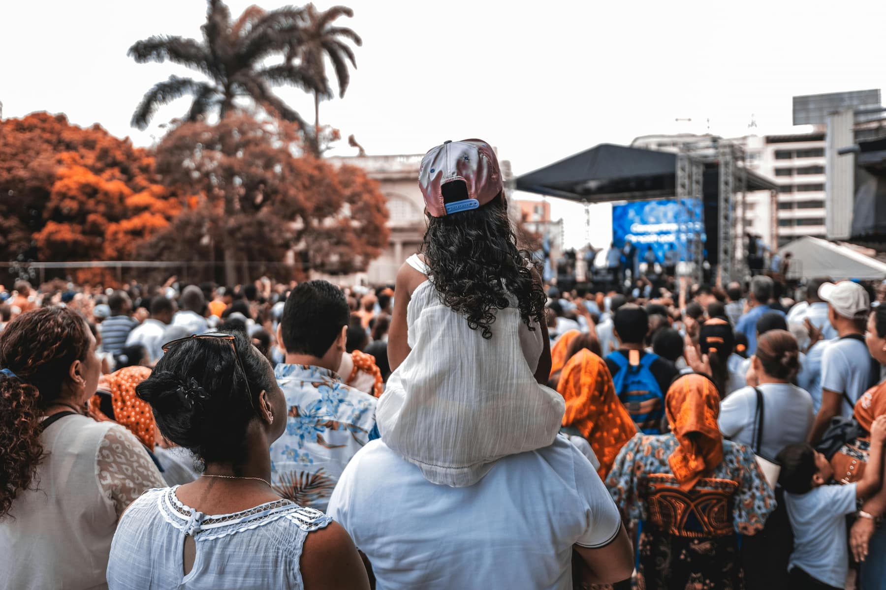 A child on someone's shoulders views a crowd and outdoor stage.