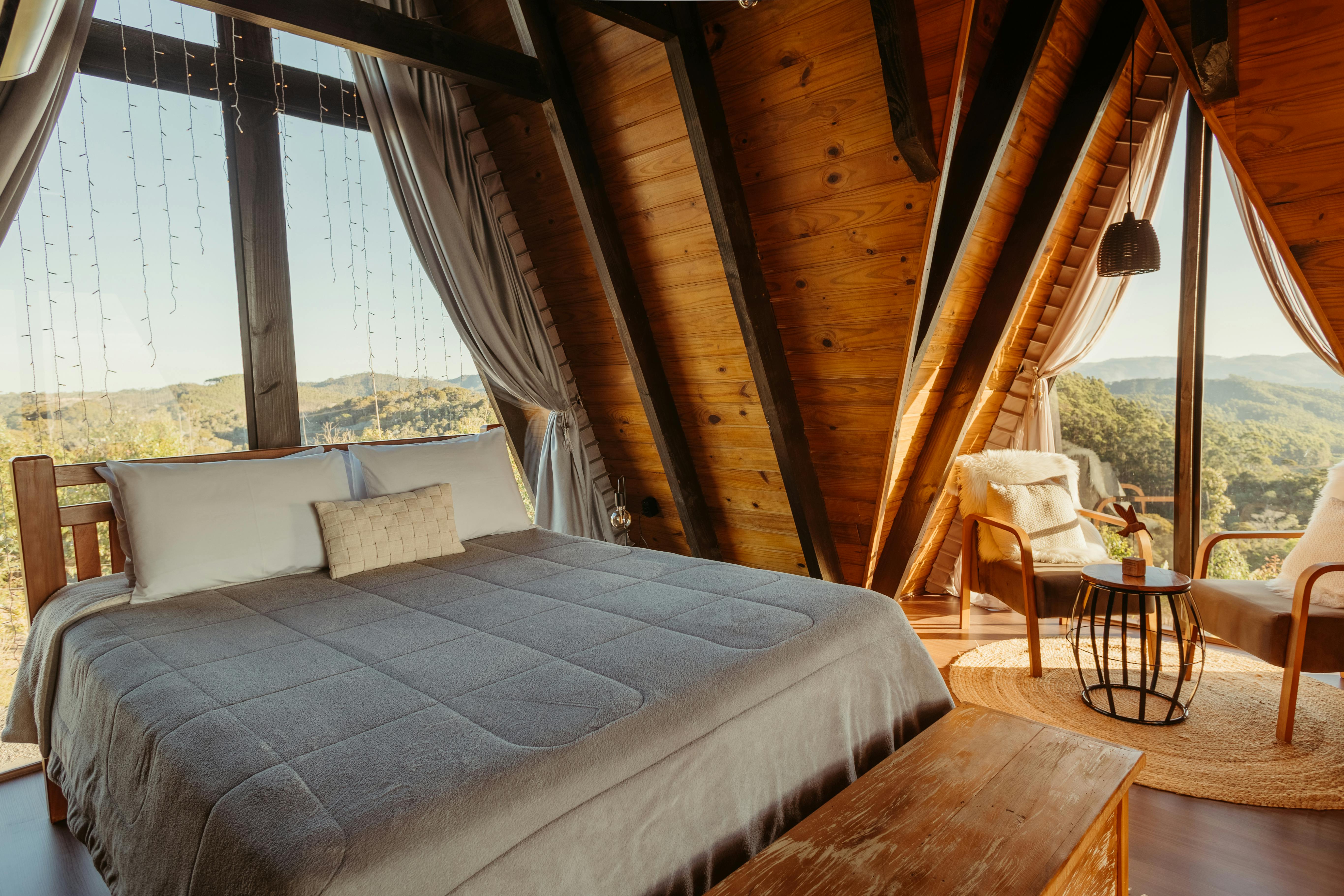 A cozy bedroom with a large bed, wooden beams, and expansive windows offering a beautiful nature view.