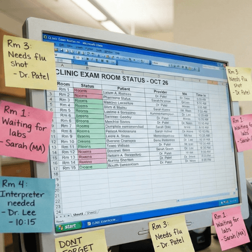 Computer screen displaying a clinic exam room status spreadsheet with sticky notes highlighting room flu shots, lab waiting, and interpreter needs.