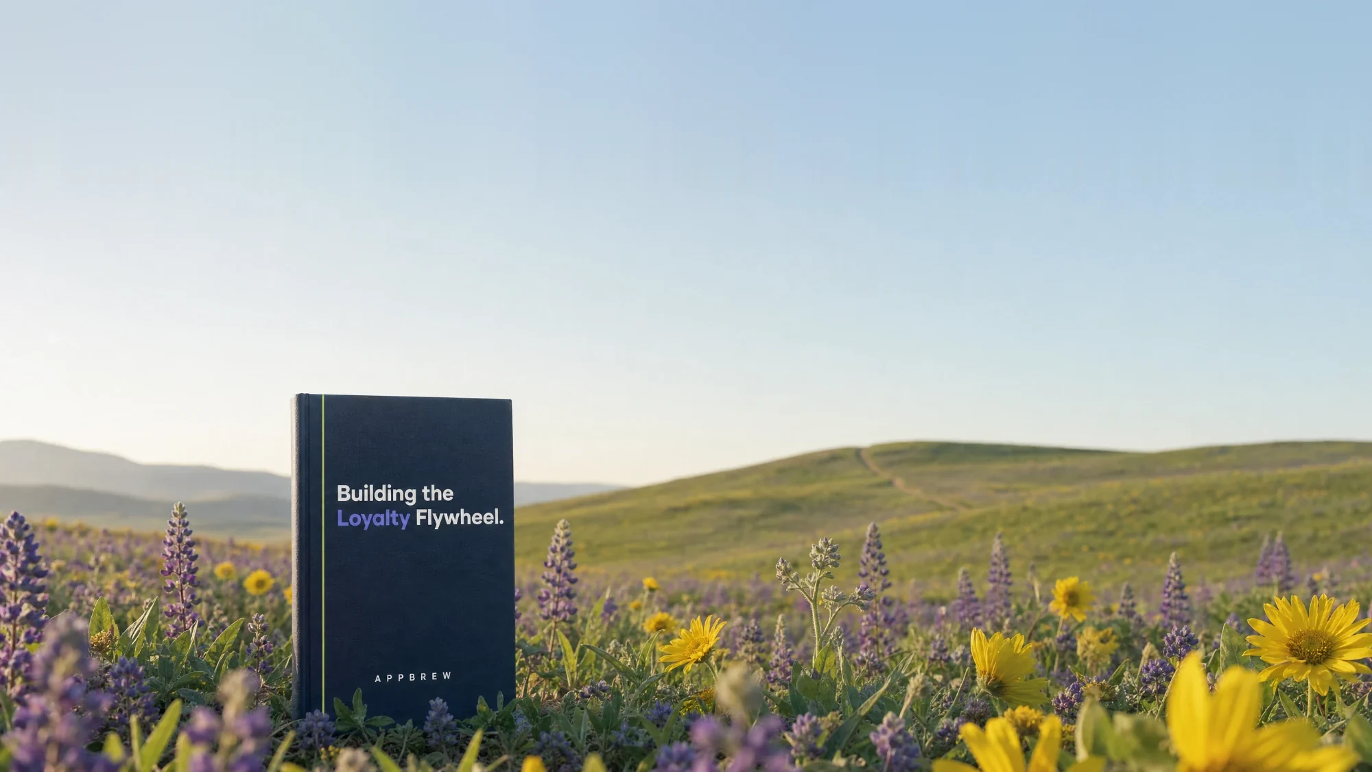 Building the Loyalty Flywheel, a navy hardcover eBook standing in a meadow of lupine and balsamroot wildflowers