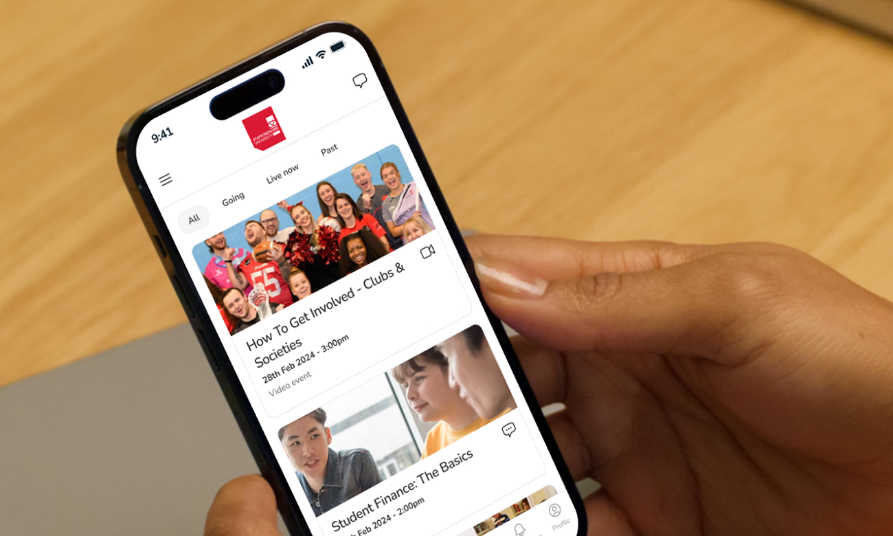 A person holding a smartphone displaying the CampusConnect app. The screen shows an events feed with options like "How To Get Involved - Clubs & Societies," scheduled for 28th February 2024, and "Student Finance: The Basics," scheduled earlier the same day. The Staffordshire University logo is visible at the top of the app interface. The background shows a wooden table and a gray object.