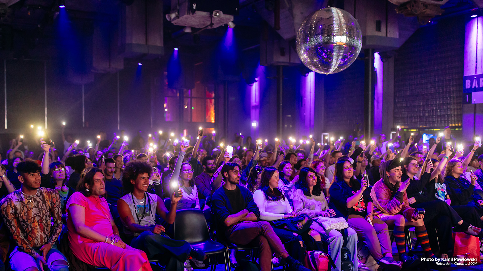 Audience at a concert venue holding up phones with flashlights on under purple lighting and a large disco ball overhead.