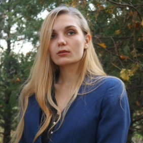 My photo. A woman with long blonde hair wearing a blue blazer stands outdoors in front of trees with green leaves.