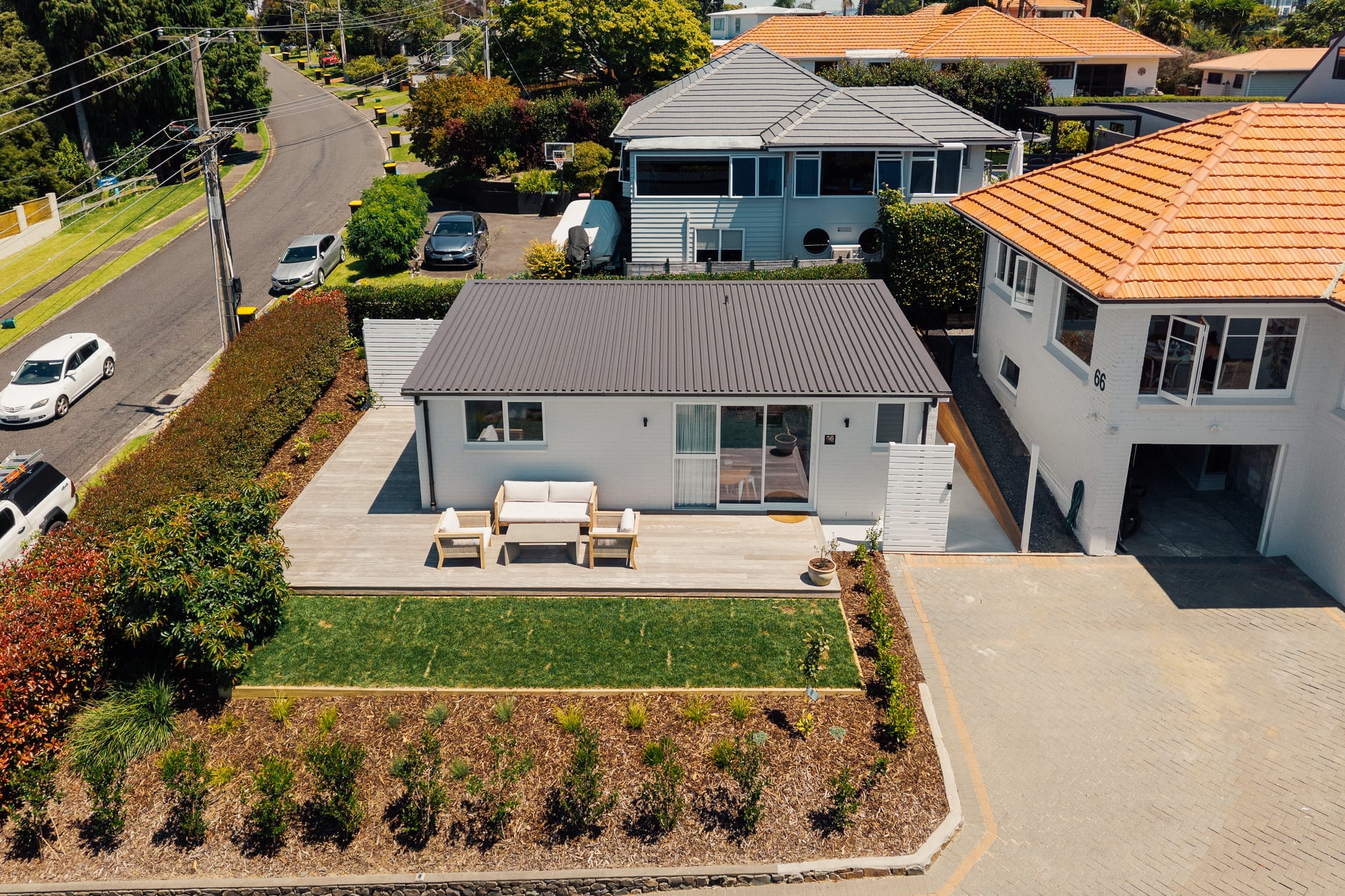 Aerial view of a modern white house with a gray roof, outdoor seating on a wooden deck, manicured lawn, and landscaped garden at the front.