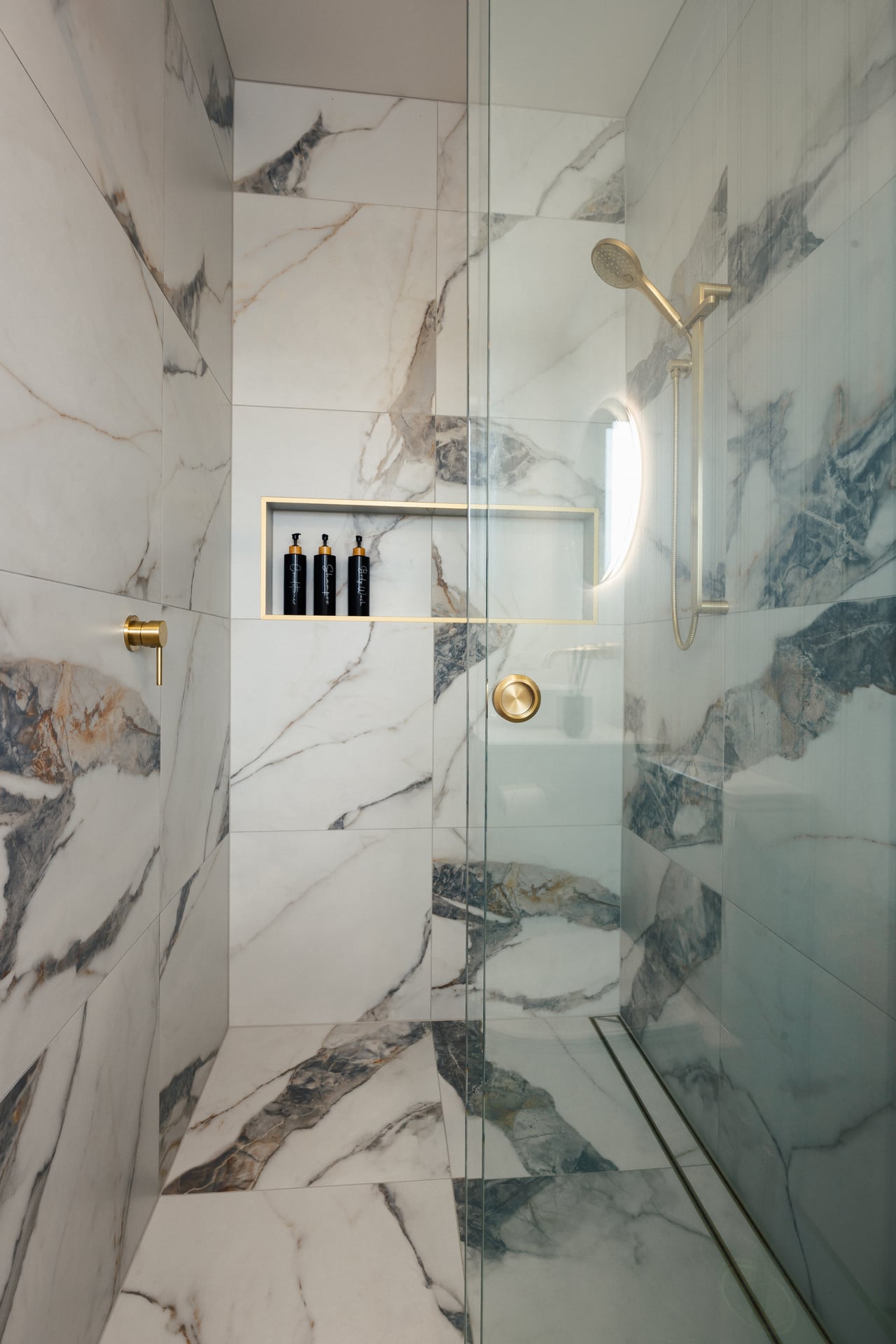 Modern shower with white and gray marble tiles, gold fixtures, and built-in shelf holding three black bottles.