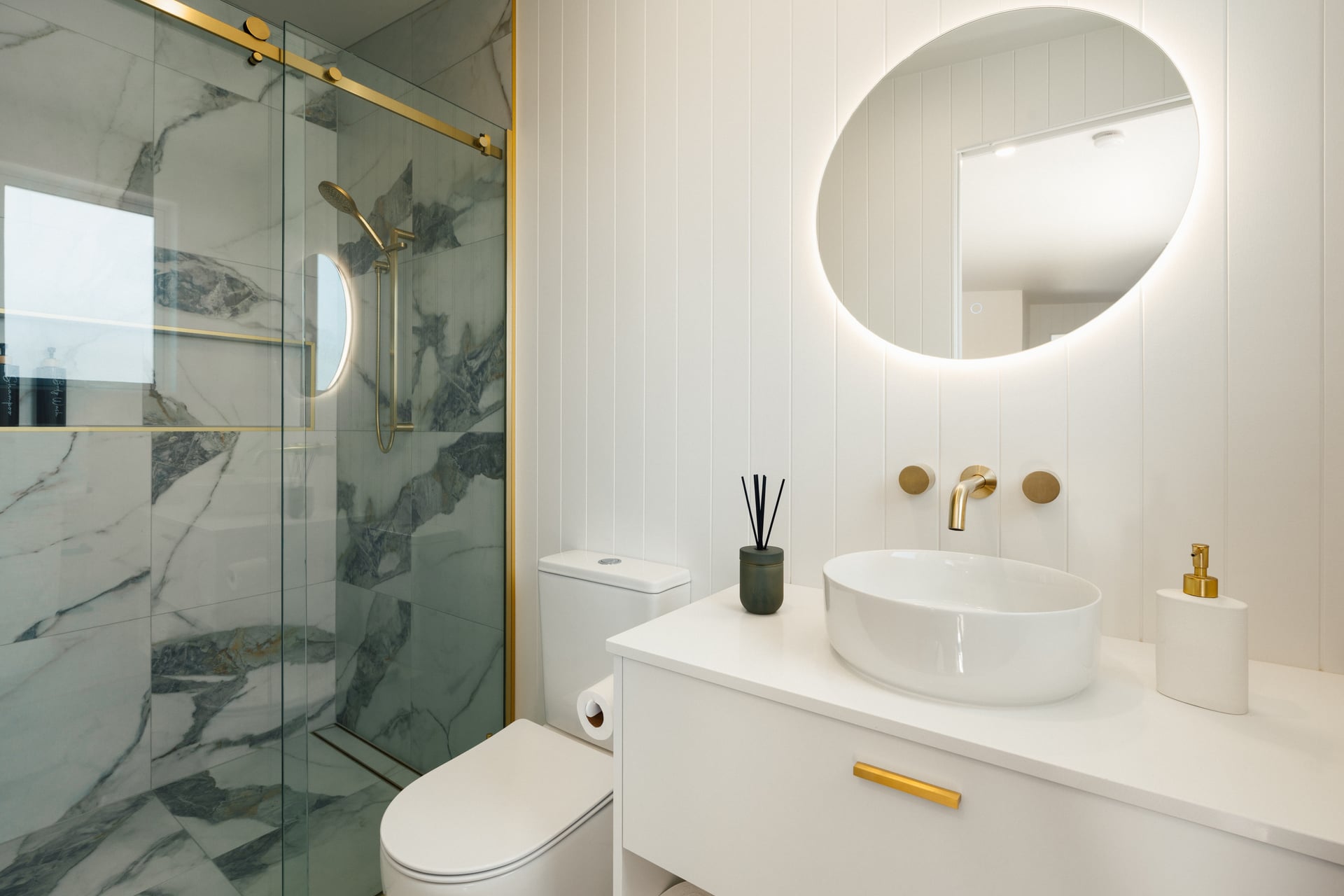 Modern bathroom with marble-tiled shower, gold fixtures, white toilet, illuminated oval mirror, and white vanity with round sink and soap dispenser.