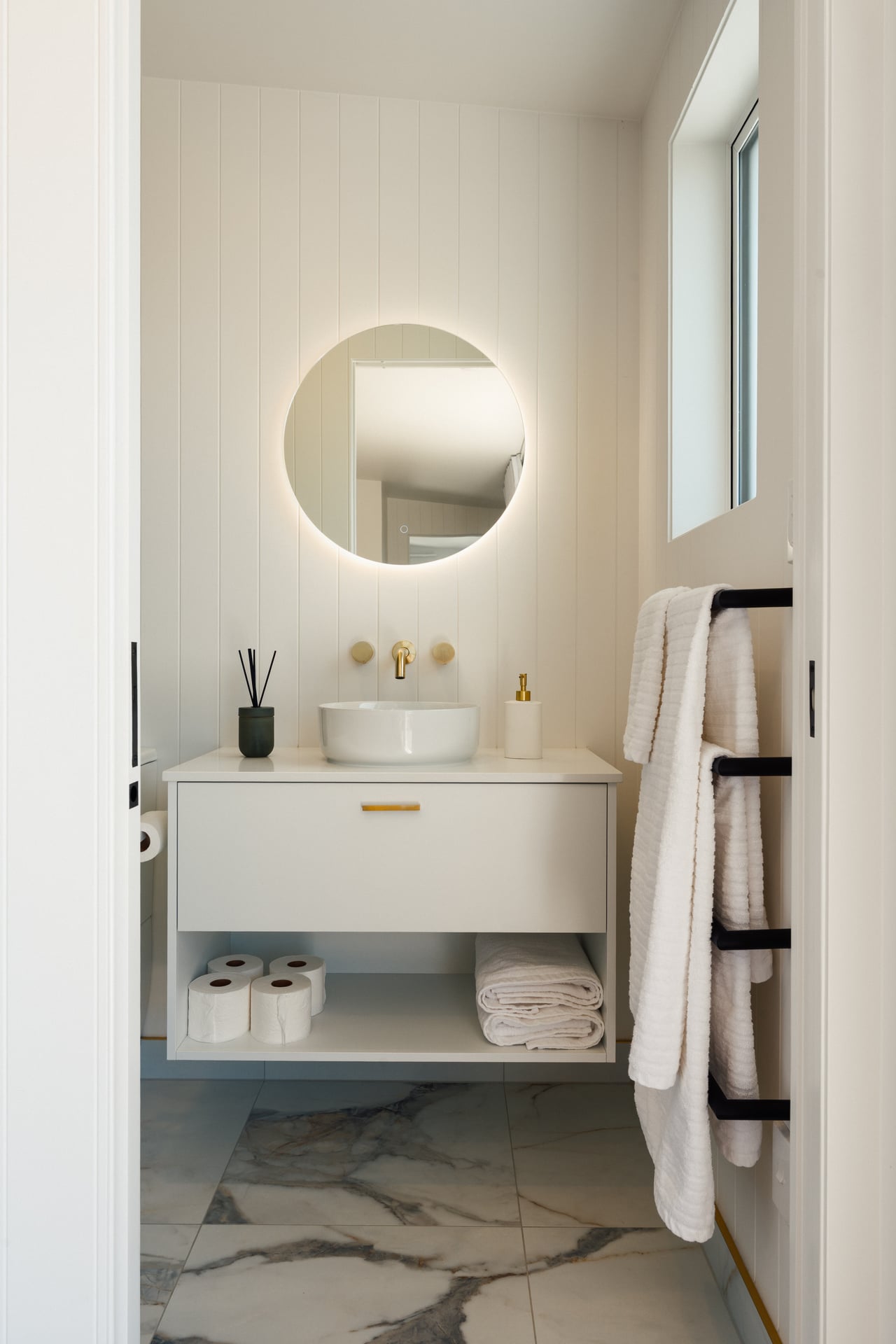 Minimalist bathroom with a round backlit mirror, white sink on a floating vanity, folded towels and toilet paper rolls below, and white towels hanging on a black rack.