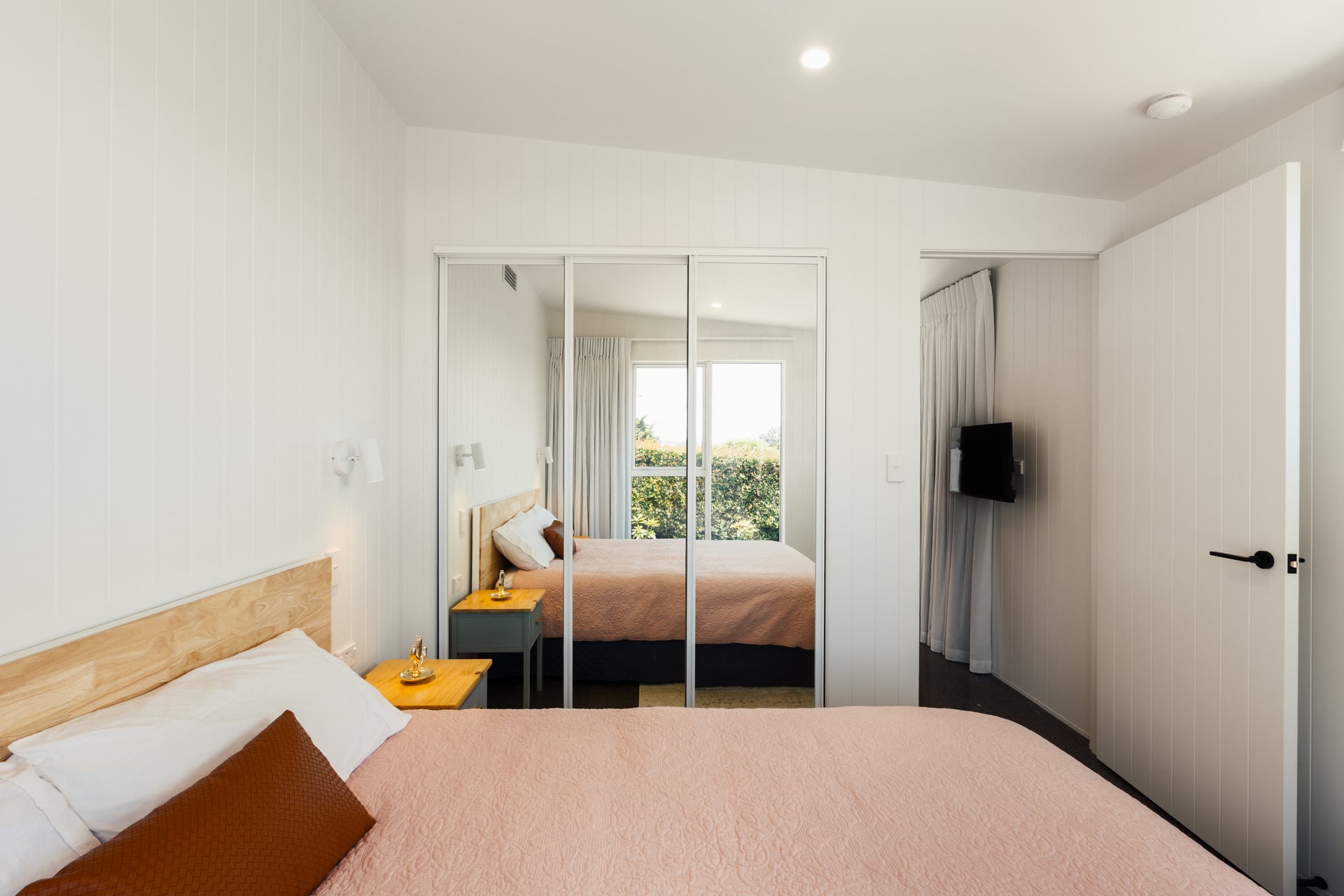 Minimalist bedroom with a wooden bed frame, pink quilt, brown pillow, nightstand with candle, mirrored closet doors, and a wall-mounted TV.