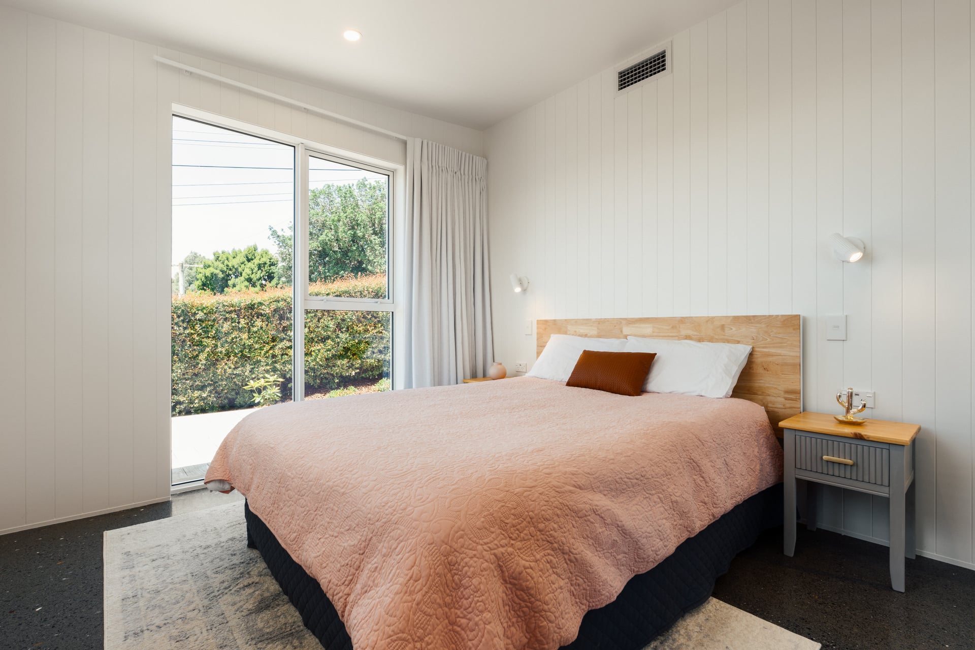 Modern bedroom with a queen bed featuring a pink quilt, white pillows, a brown accent pillow, and a wooden headboard, next to a gray nightstand and large window with white curtains.