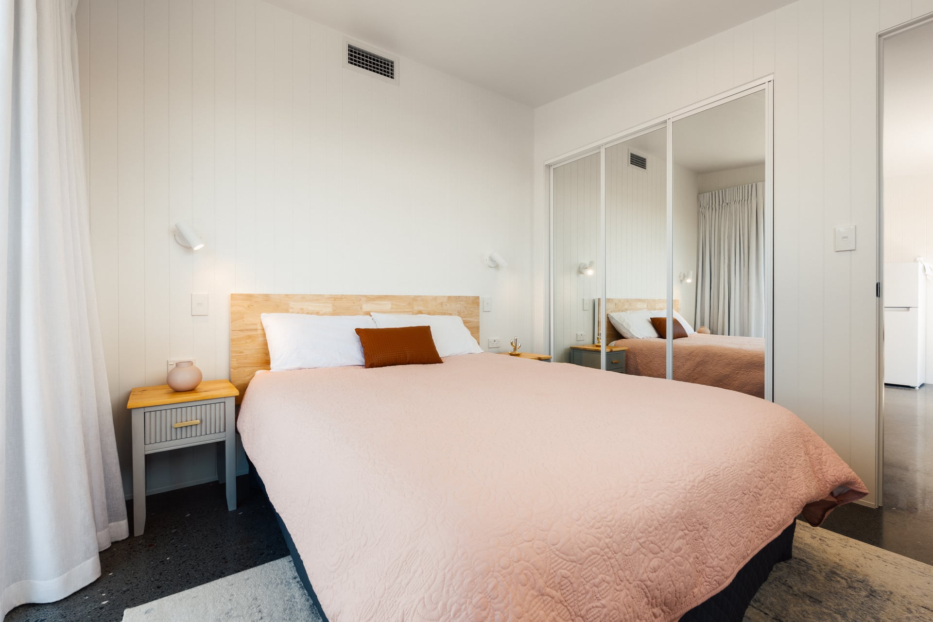 Minimalist bedroom with a bed featuring a light pink quilt, two pillows with a brown accent pillow, wooden headboard, bedside table with vase, and mirrored closet doors.