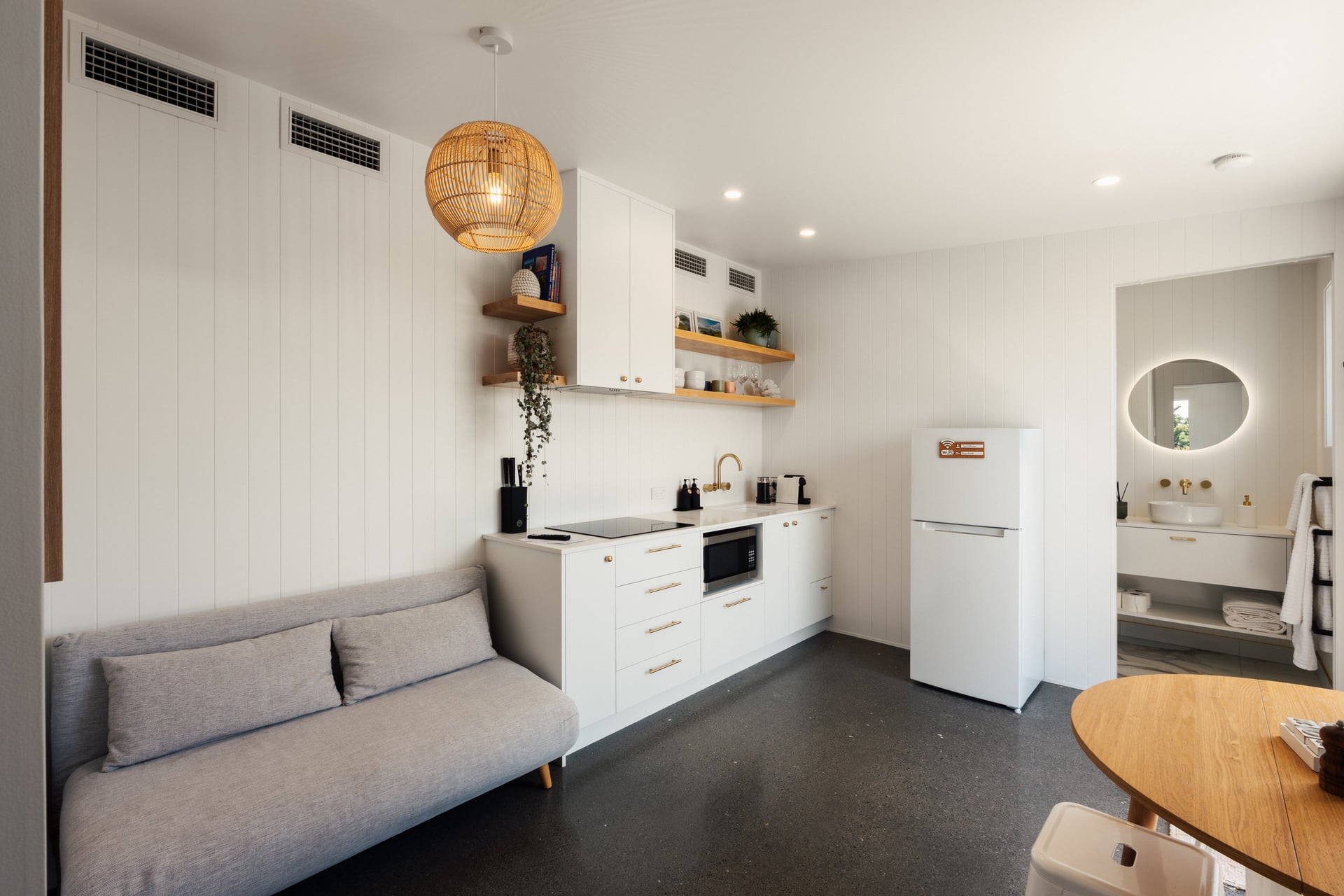 Modern kitchen and living space with white cabinets, black countertop appliances, gray sofa, wood dining table, and a round mirror in adjacent bathroom.