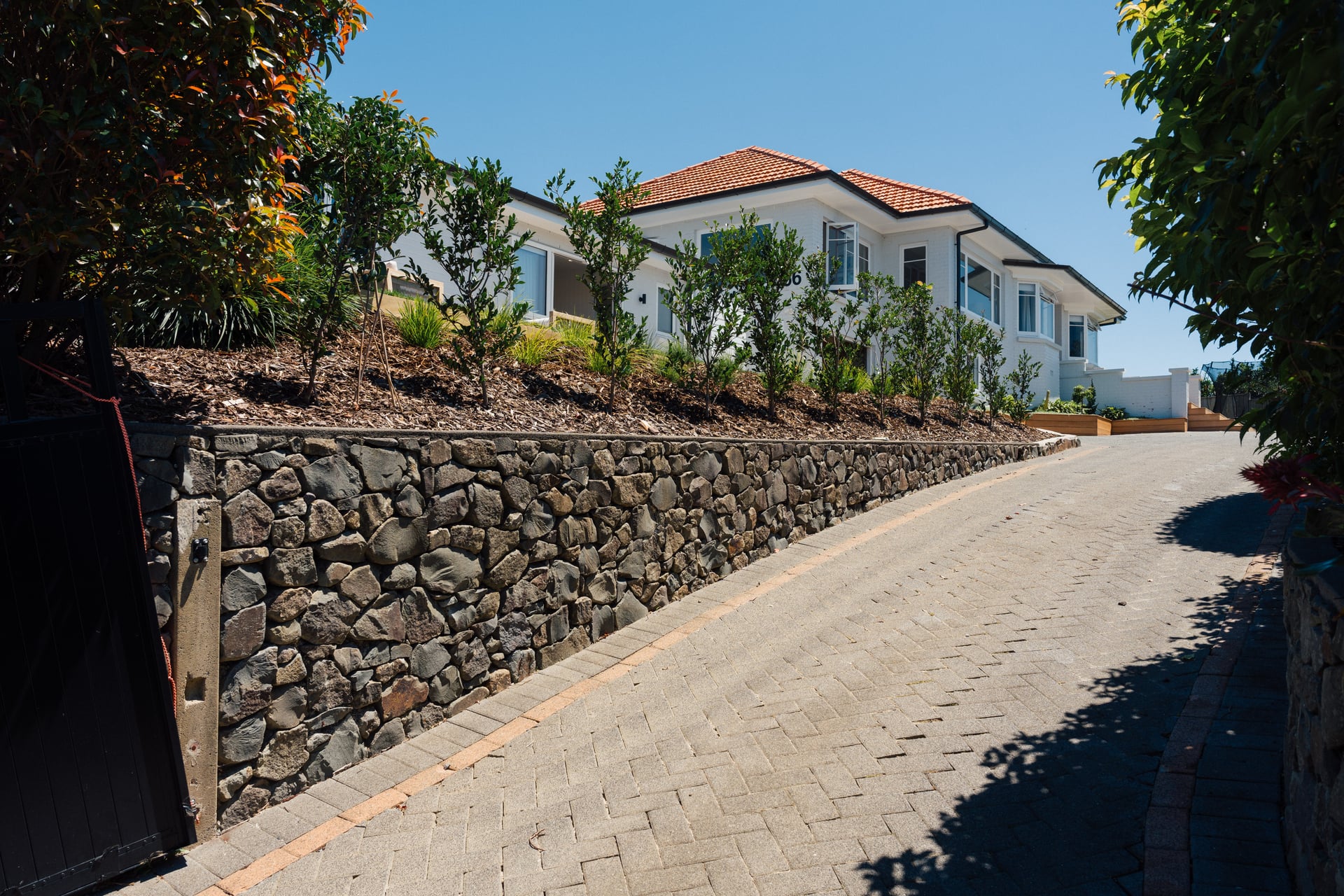 Stone retaining wall and paved driveway leading to Judea minor dwelling by Gardo Group with terracotta roof and landscaped grounds