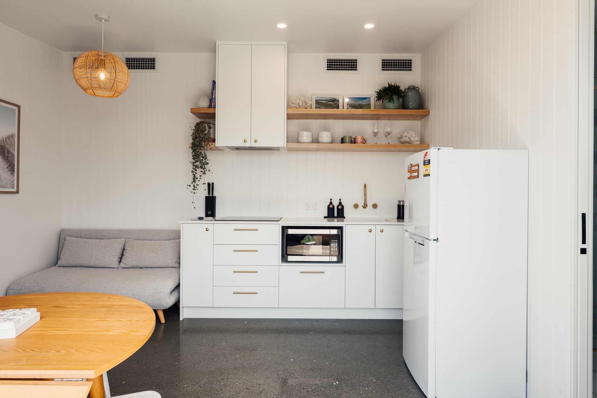 Modern kitchen with white cabinets, built-in microwave, white refrigerator, wooden shelves with plants and dishes, and a small gray sofa under a wicker pendant light.
