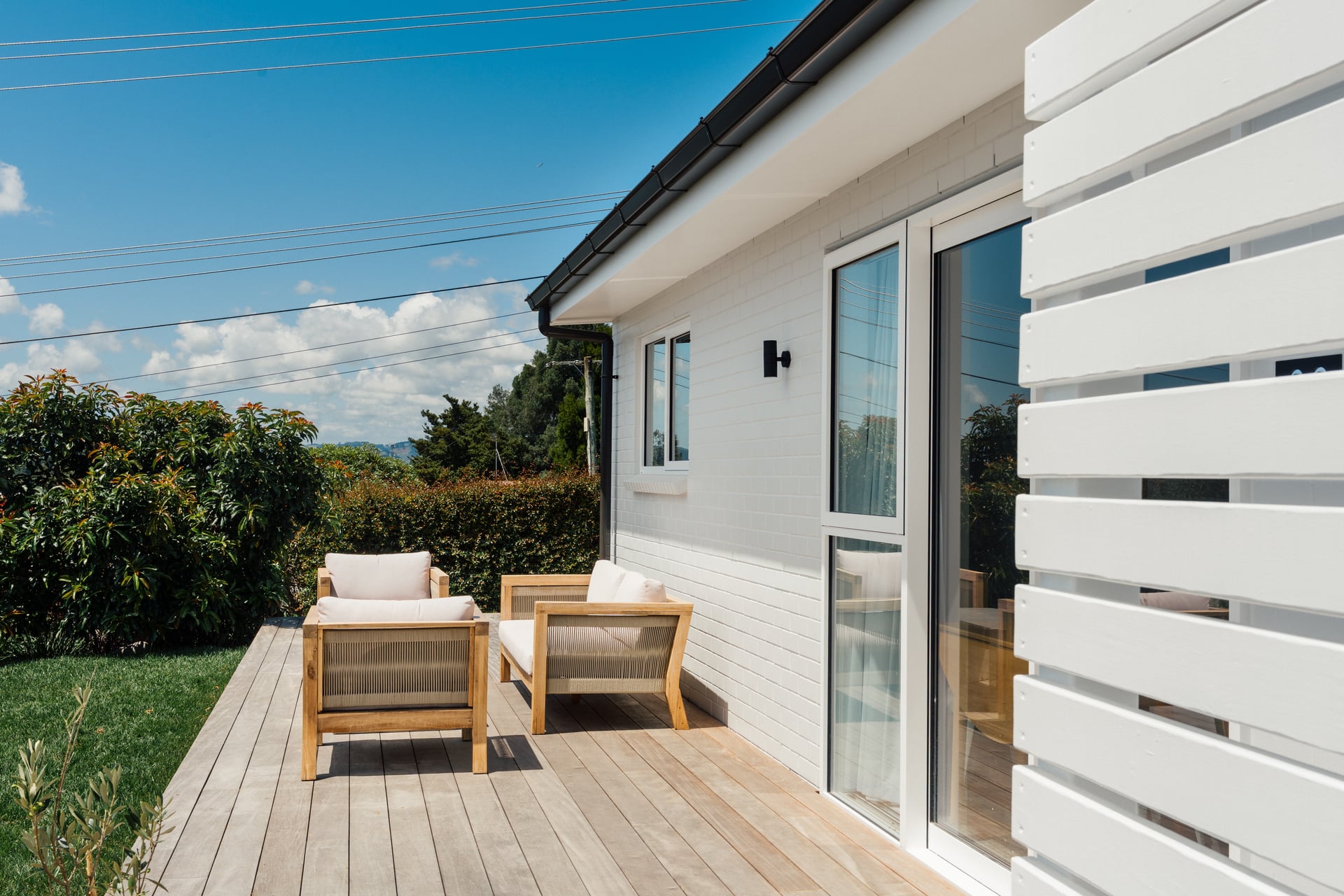 Outdoor timber deck with lounge seating at Judea minor dwelling by Gardo Group featuring white weatherboard cladding and garden views