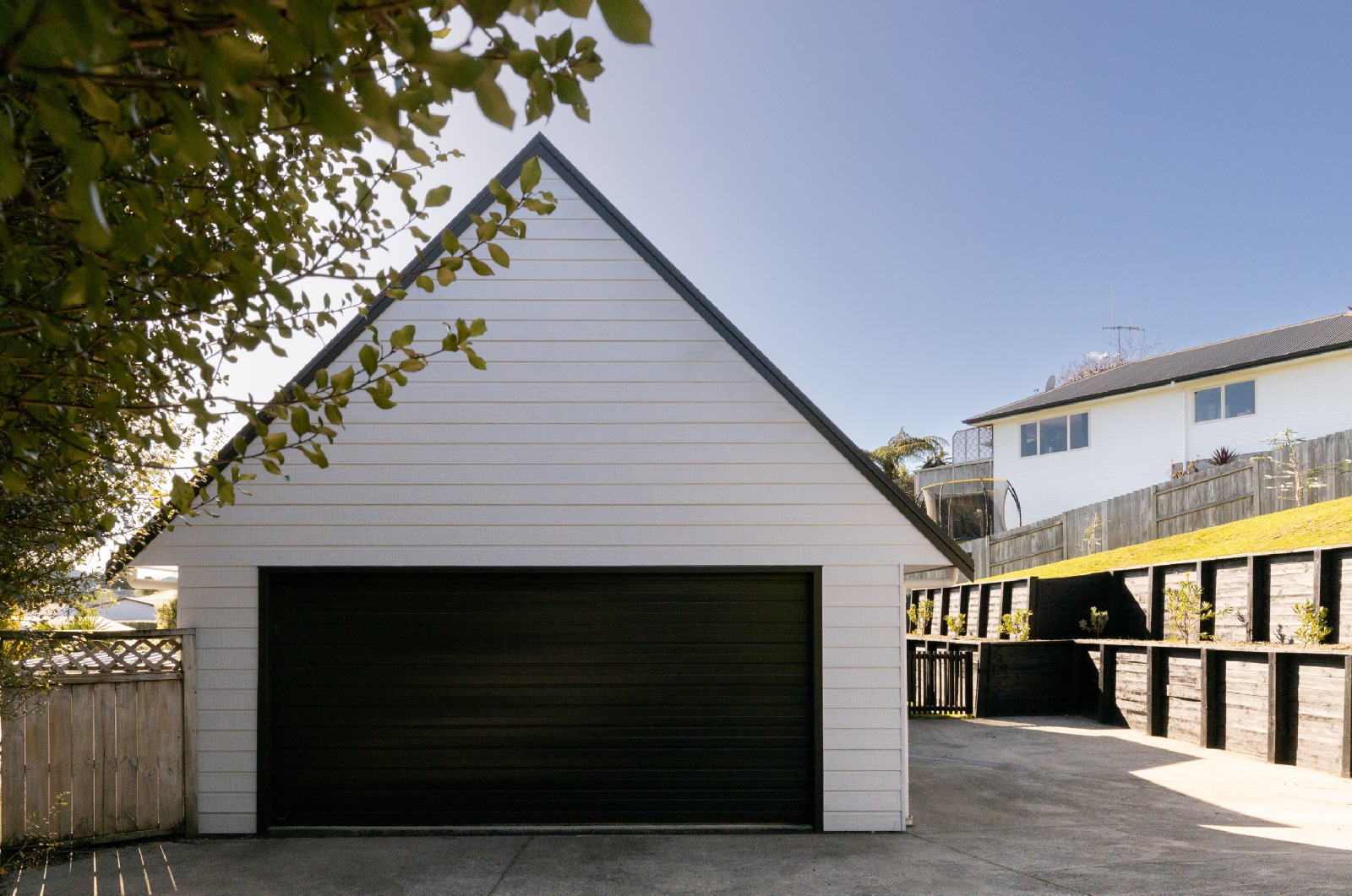 White weatherboard gable garage with a black door and timber retaining wall, built by Gardo Group at Welcome Bay