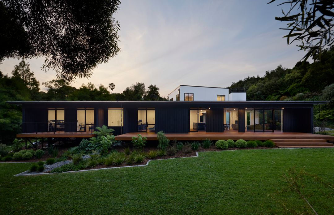 Dark vertical board clad single storey home with a full width timber deck and lit interiors at dusk, built by Gardo Group in Ōmokoroa
