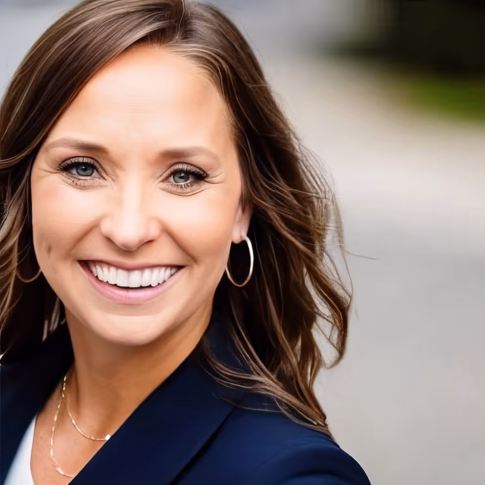 Smiling woman with light brown hair, wearing hoop earrings and a navy blazer.